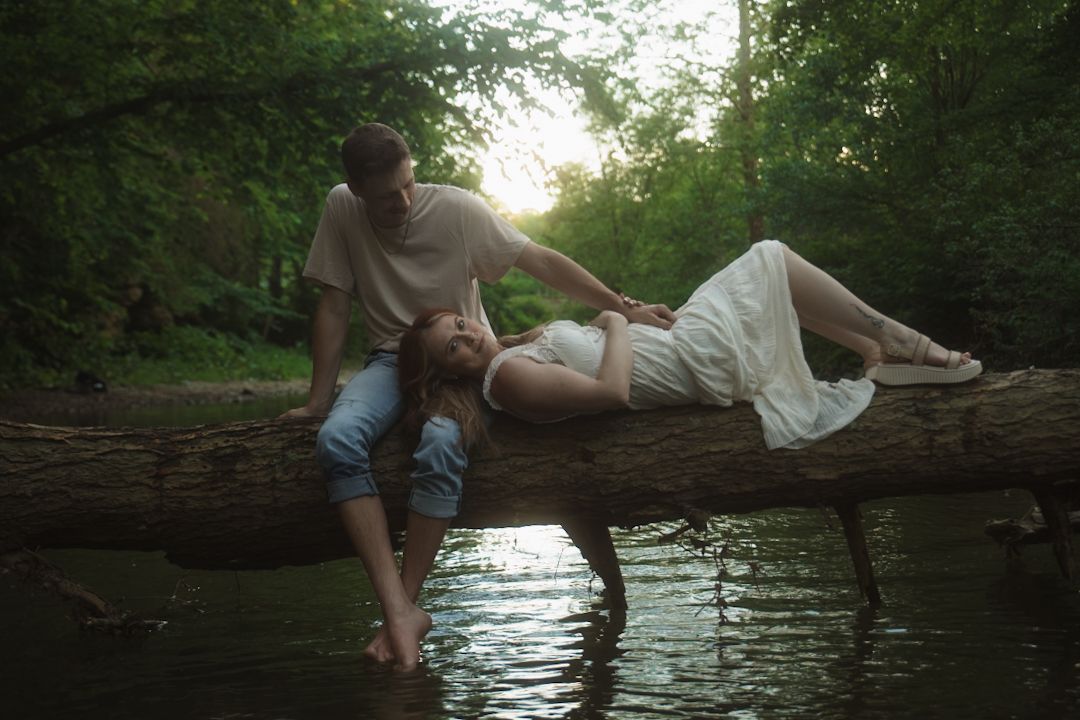 A couple rests on a fallen log over a river in a lush green forest, woman in white dress lying across the log.