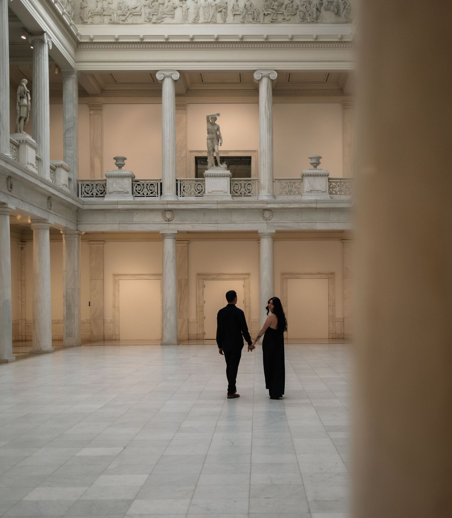 Couple holding hands in a grand classical museum atrium with marble columns.