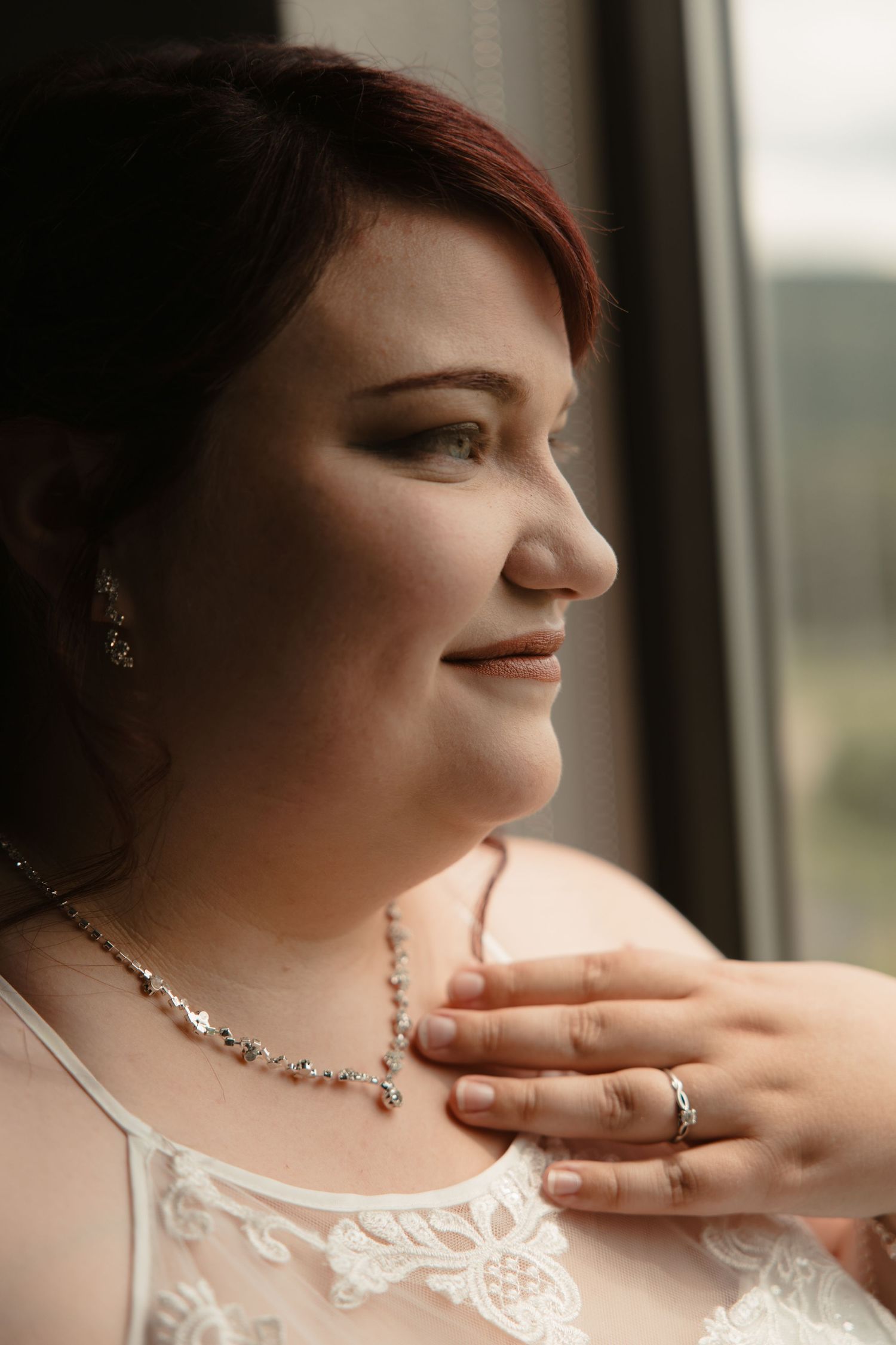 A smiling bride in a white lace dress gazes out a window, wearing a delicate necklace and earrings on her wedding day.