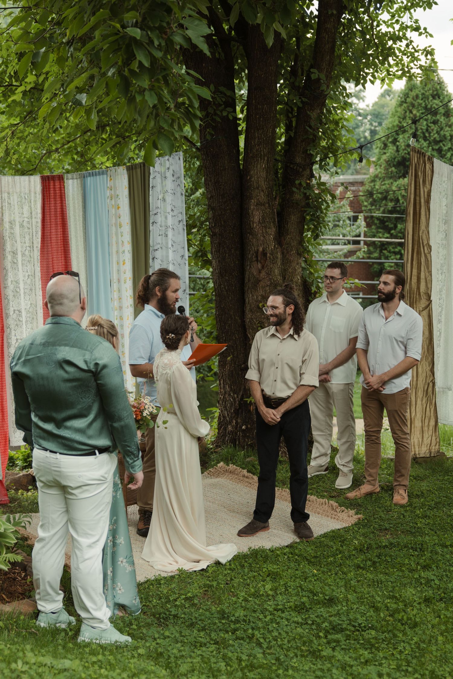 Outdoor wedding ceremony in garden with colorful fabric backdrop, bride in white dress, groom, officiant, and groomsmen on green lawn.