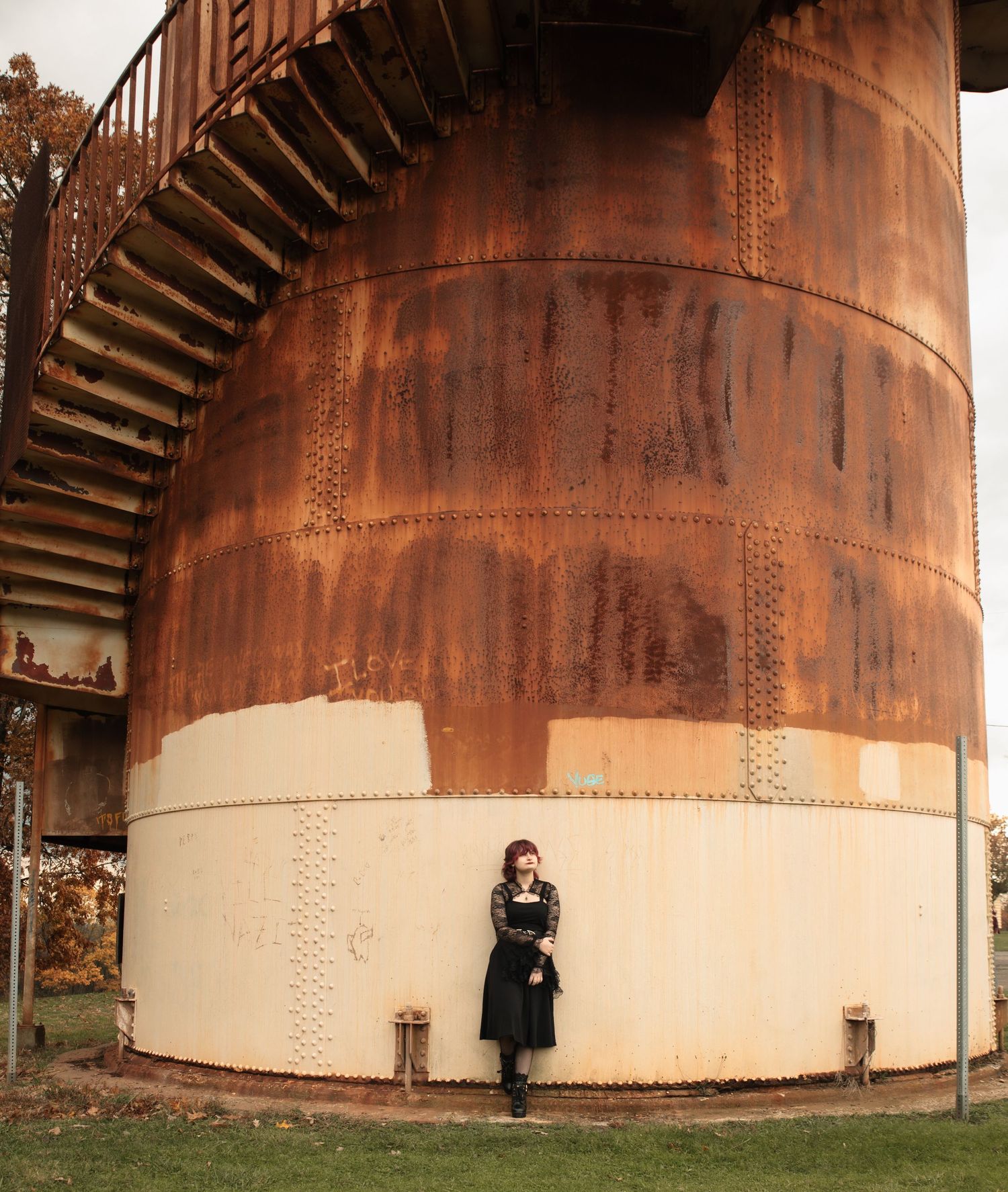 A person stands against a large rusted industrial storage tank showing its massive scale and weathered metal surface.