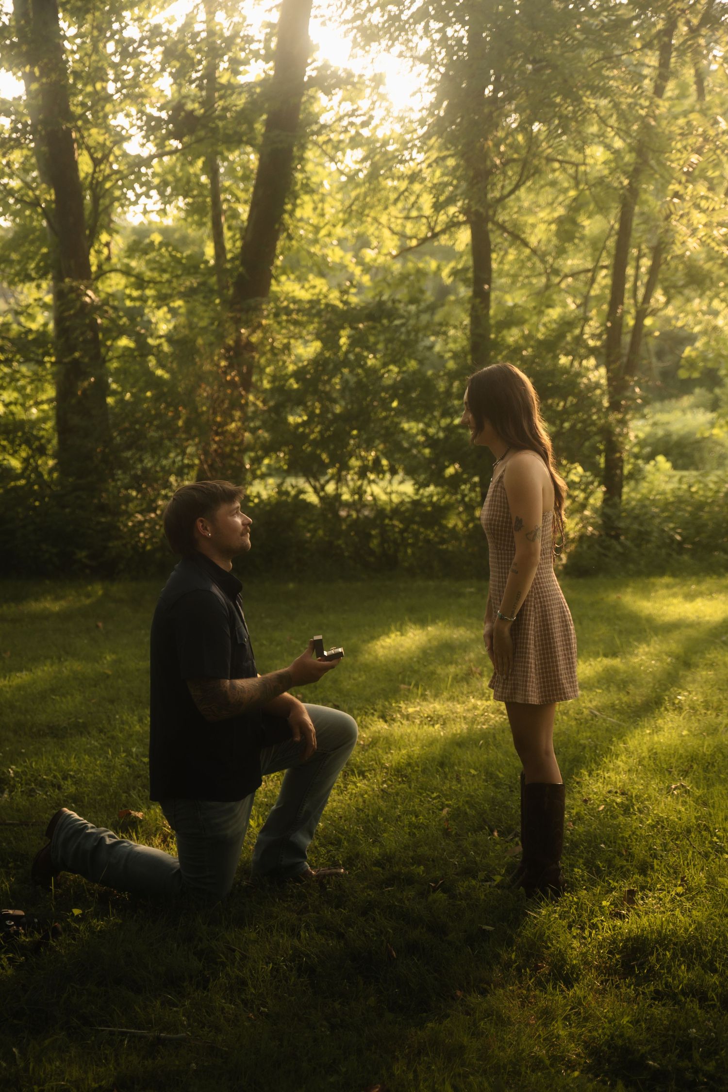 Man kneeling on one knee proposing to woman in sunlit forest clearing, holding ring box in romantic outdoor marriage proposal.