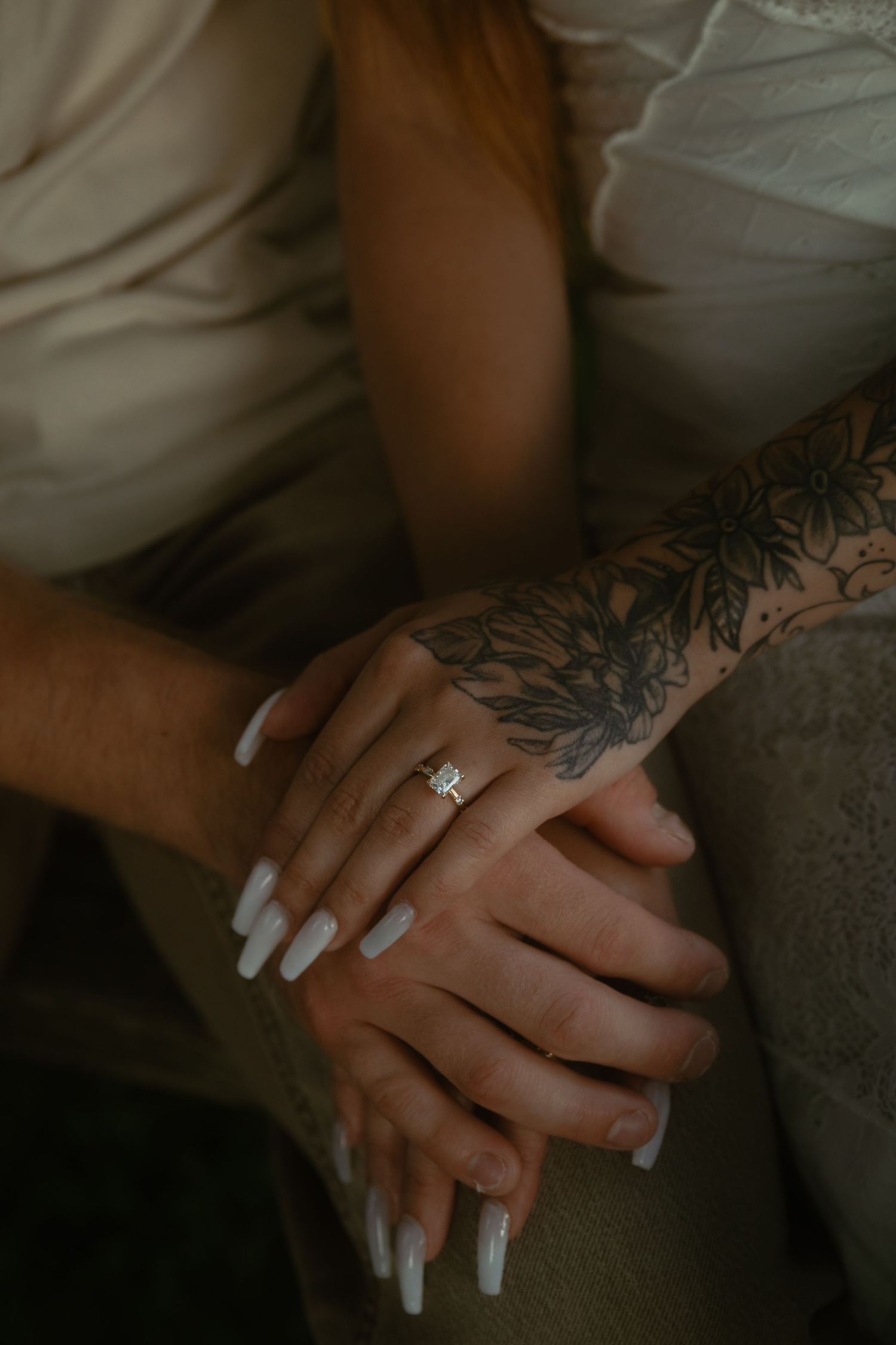Close-up of a woman's hand with white nails and an emerald-cut engagement ring resting on a man's hand, her arm tattooed.