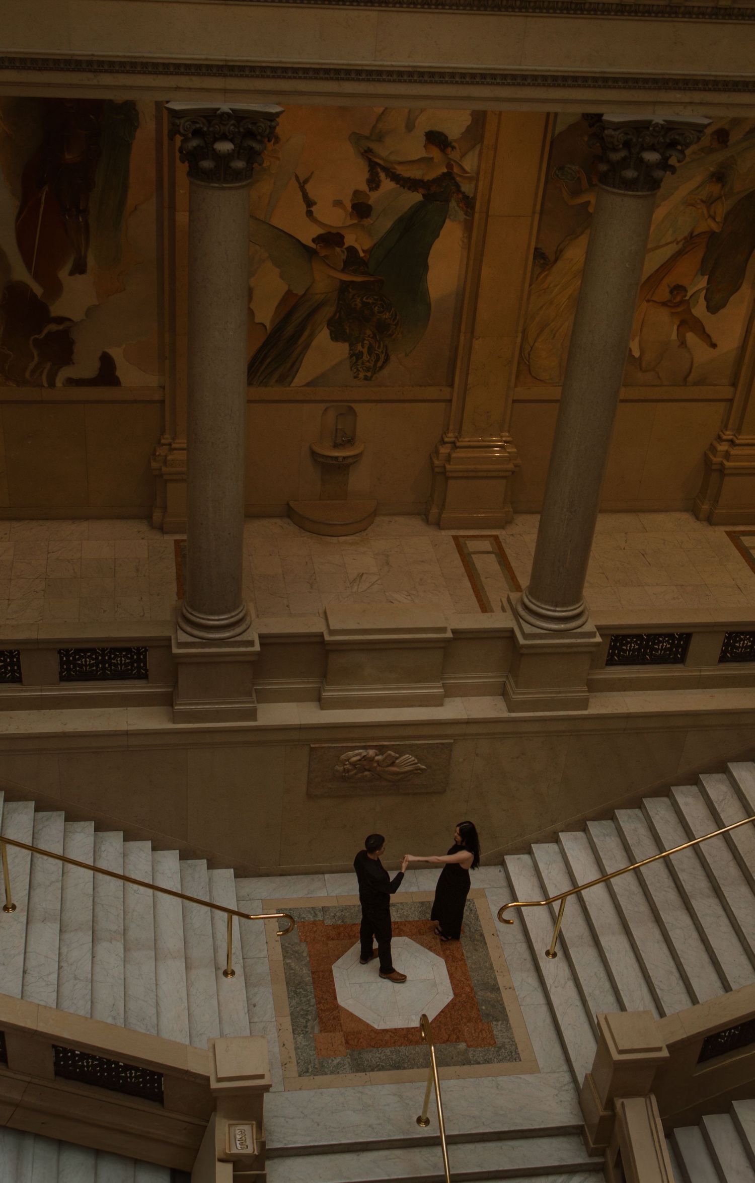 Aerial view of couple holding hands on ornate staircase landing in grand classical building with murals.