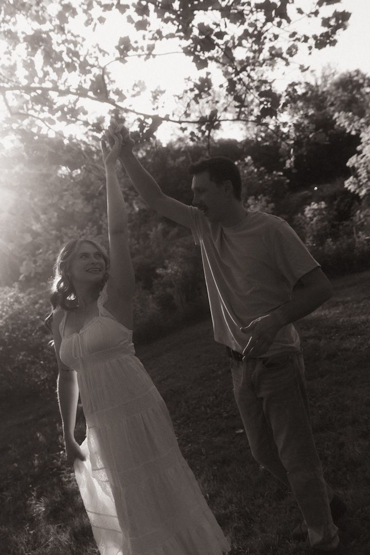 Black and white photo of a romantic couple dancing outdoors, woman in white dress, man spinning her under trees in sunlight.