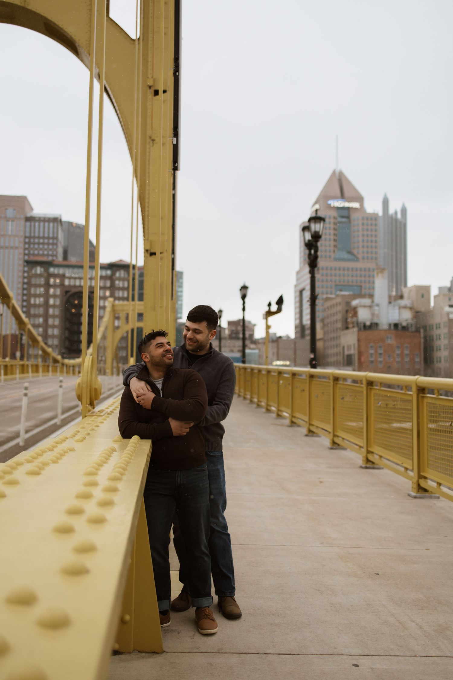 Two men embrace on a yellow bridge with Pittsburgh city skyline in the background on a cloudy day.