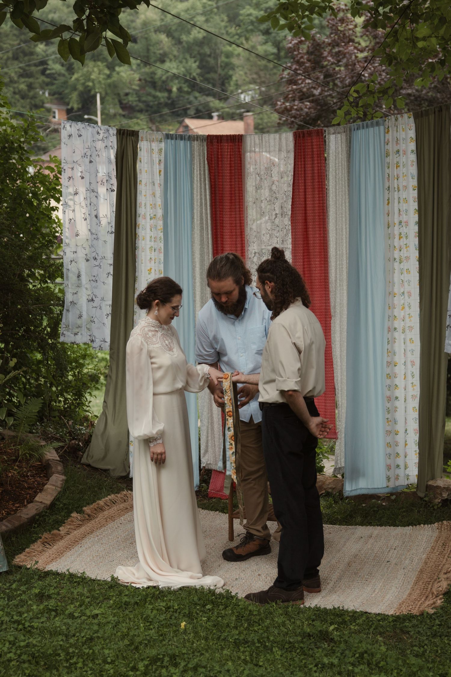 Outdoor wedding ceremony with bride in white gown, officiant, and groom before colorful fabric backdrop on green lawn.
