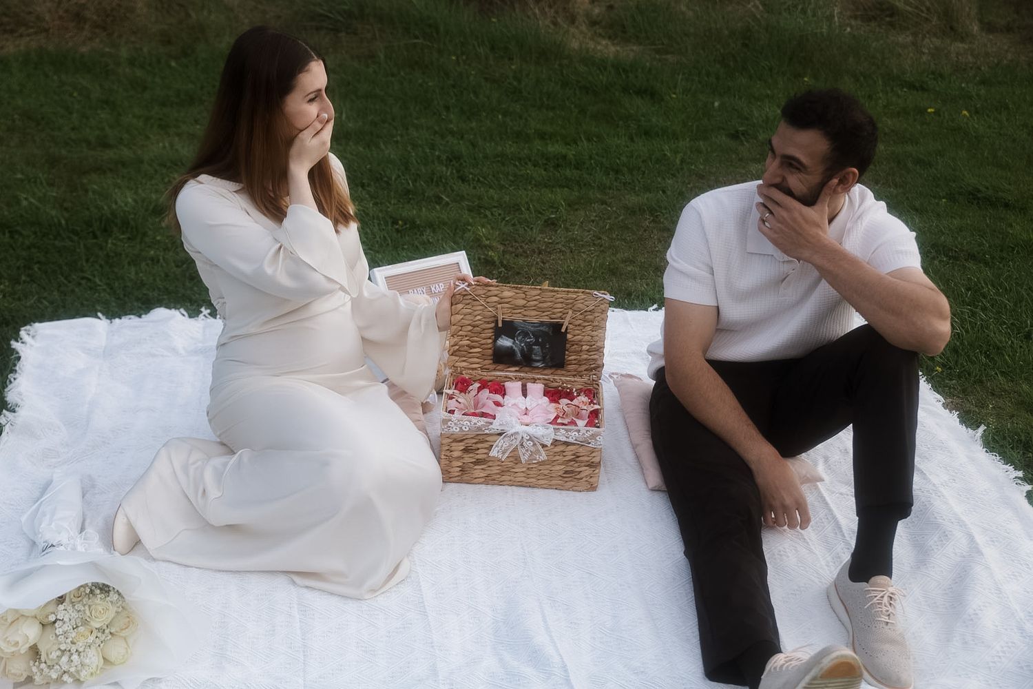 Pregnant couple sitting on white blanket outdoors, looking at ultrasound photo with a decorative basket between them.