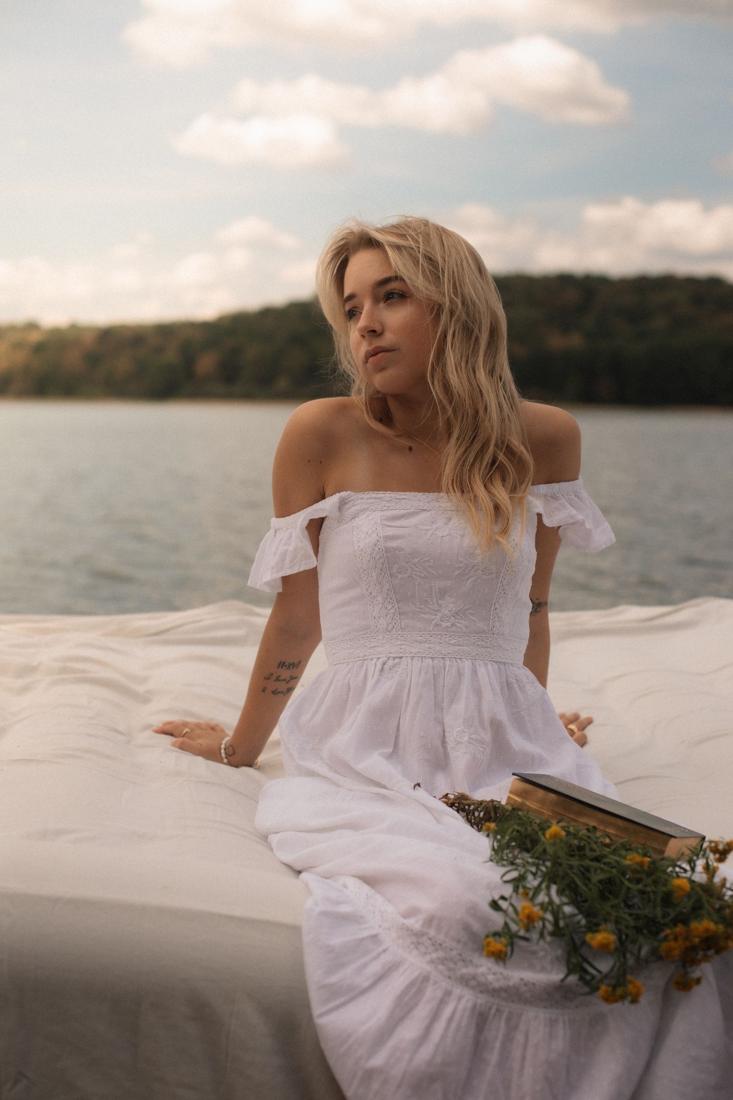 Blonde woman in white off-shoulder dress sitting outdoors near a lake, holding yellow flowers with trees in background.
