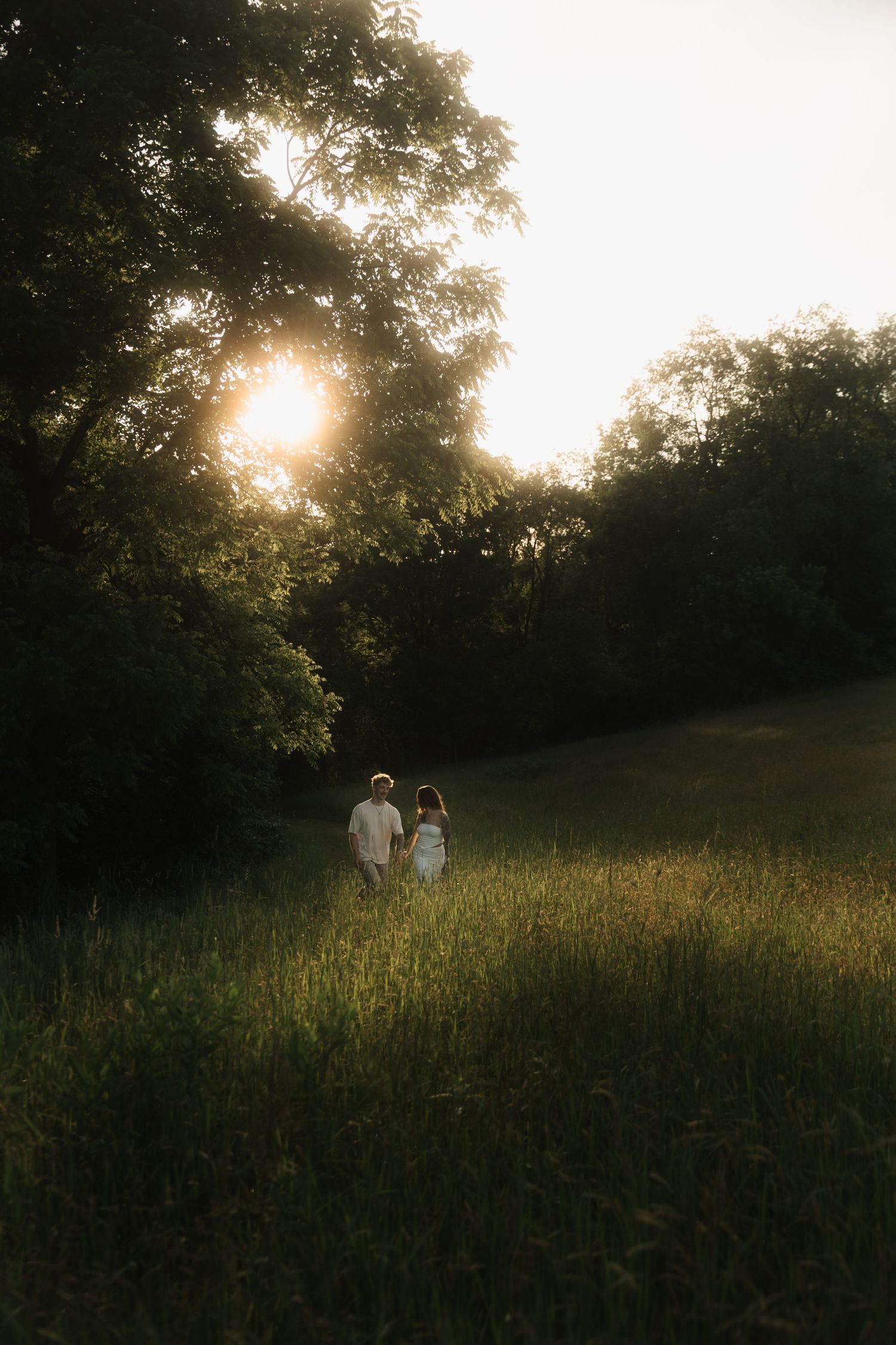 Couple holding hands walking through lush green field toward glowing sunset through trees.