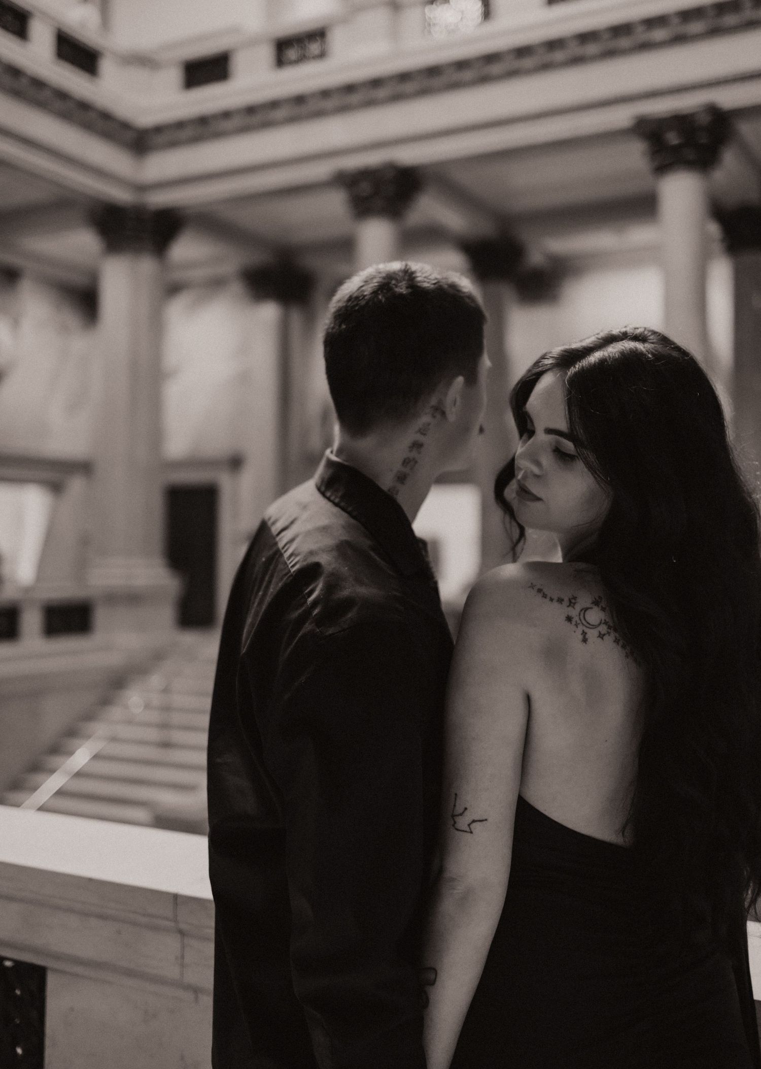Black and white photo of a couple sharing an intimate moment in an elegant columned building, woman with back tattoos.