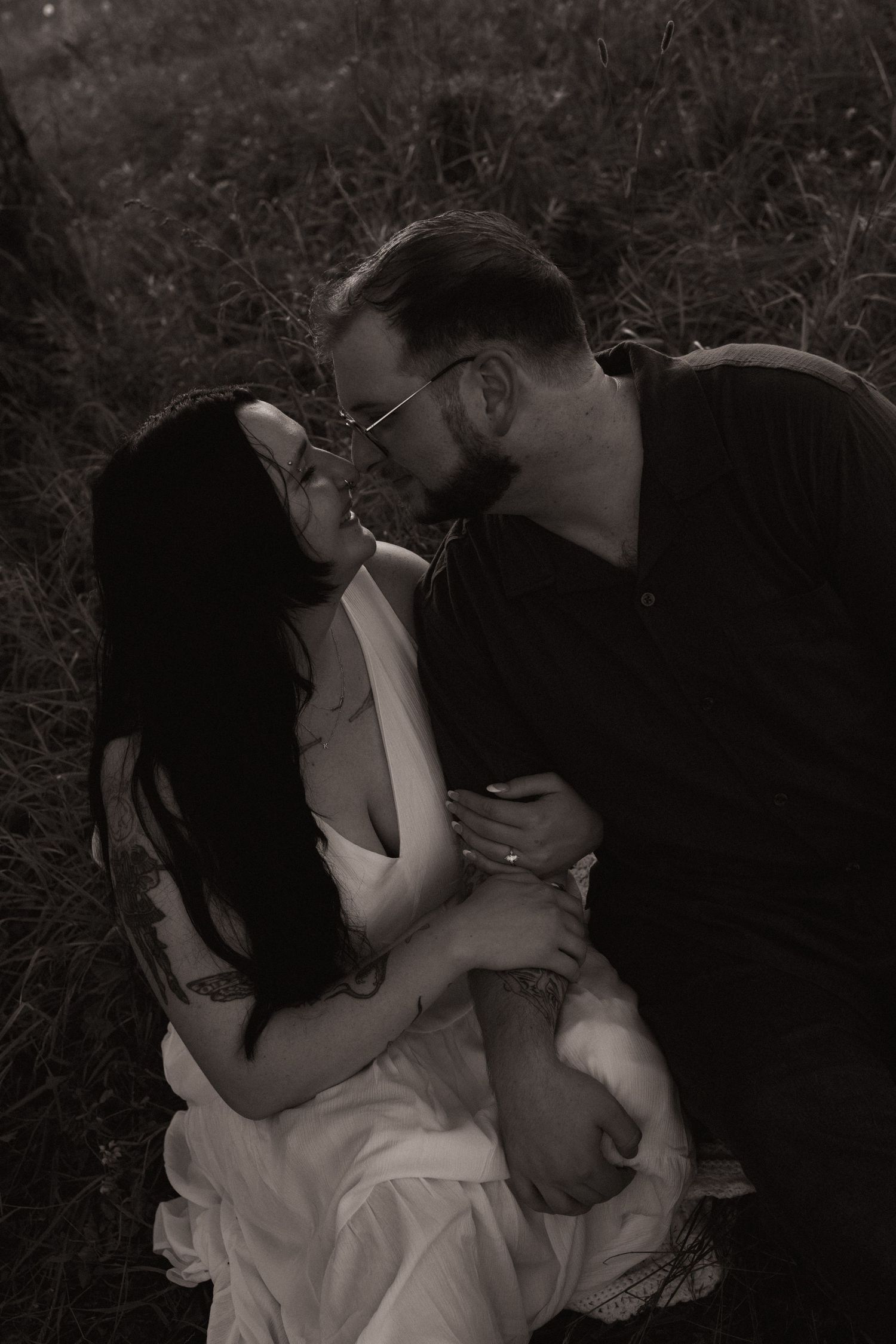 Black and white photo of a couple sharing a romantic moment, leaning in for a kiss outdoors in a grassy field.