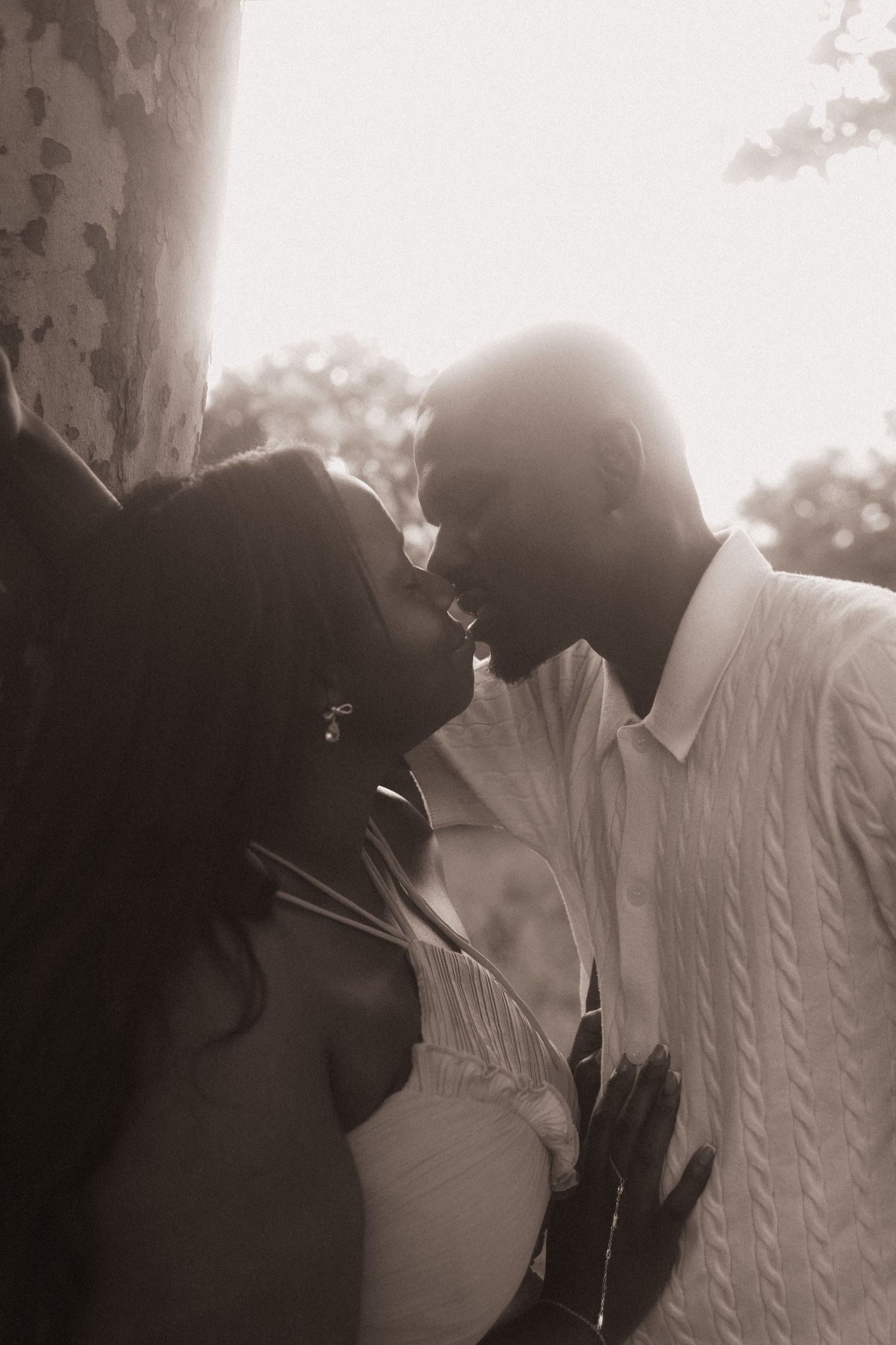 A romantic black and white photo of a couple sharing an intimate moment near a tree in soft backlit sunlight.