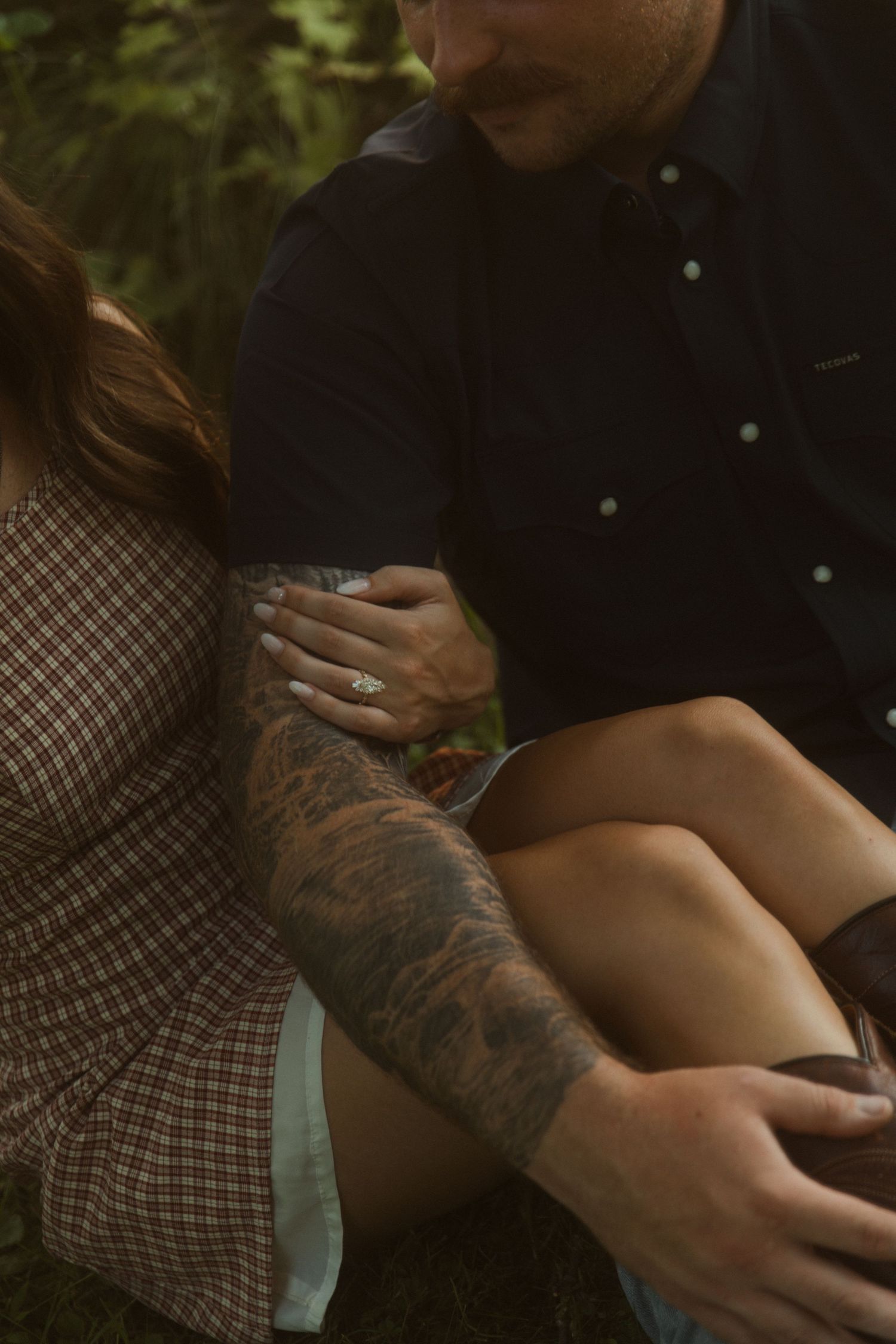 Couple sitting closely together outdoors, woman with floral tattoo sleeve wearing engagement ring resting hand on man's leg.