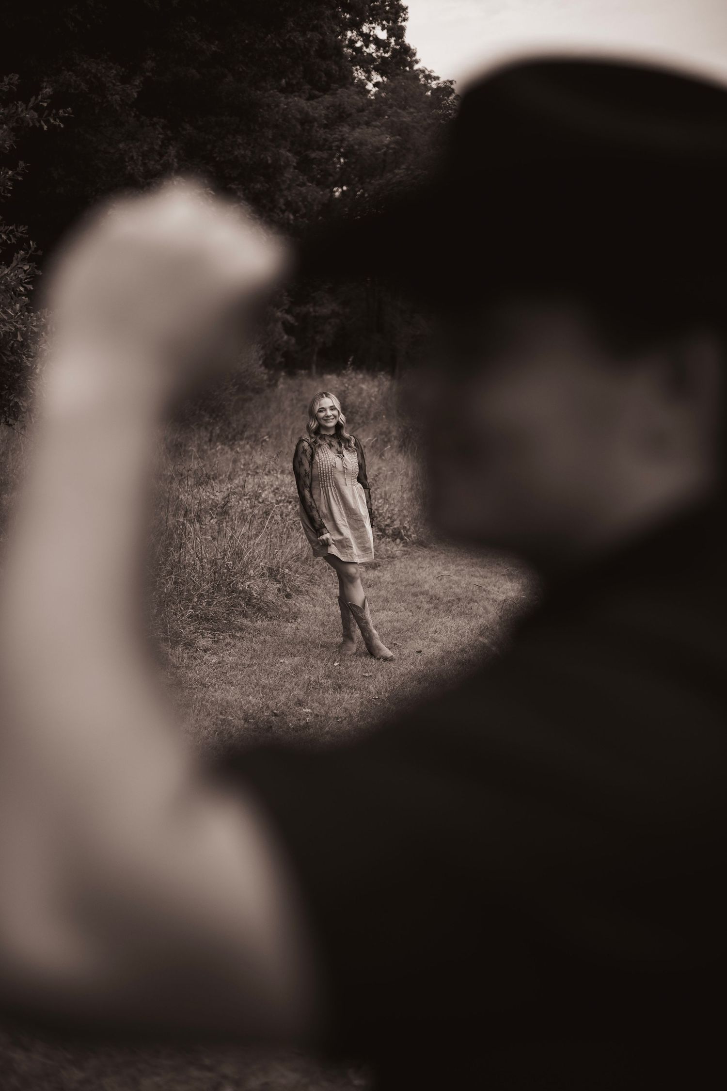 Black and white photo of a woman standing in a field framed by two blurred figures in the foreground.