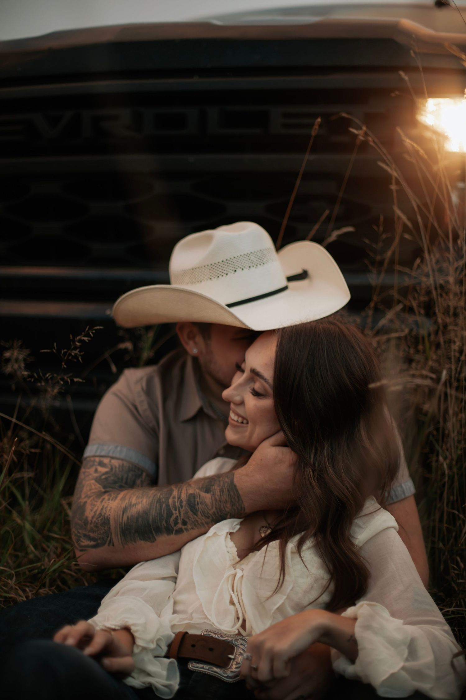 A romantic photoshoot at sunset showing a couple in western attire sharing tender moments in a field.