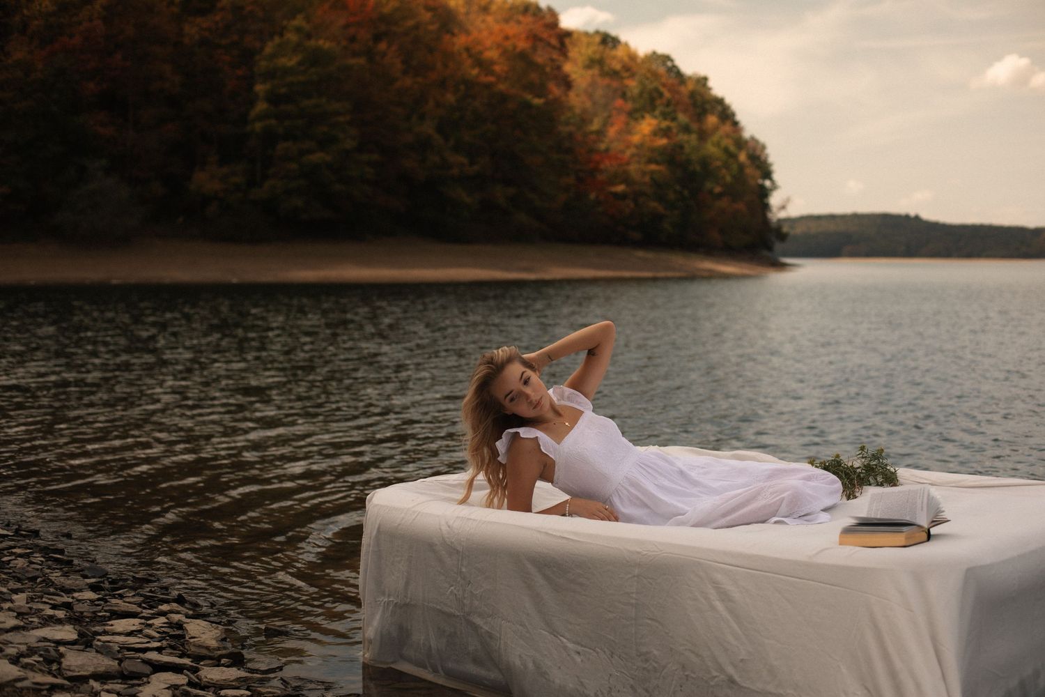 A couple lounges on a white mattress floating on a serene lake during sunset.