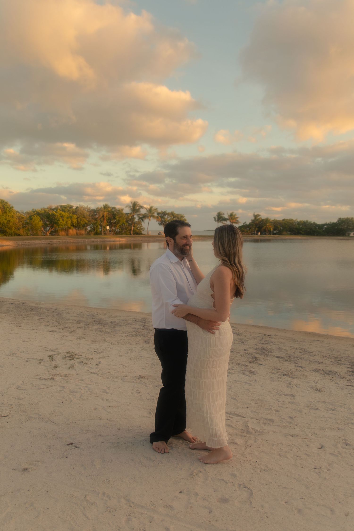 A couple shares an intimate moment at sunset on a sandy beach beside a tranquil lake with golden clouds above.