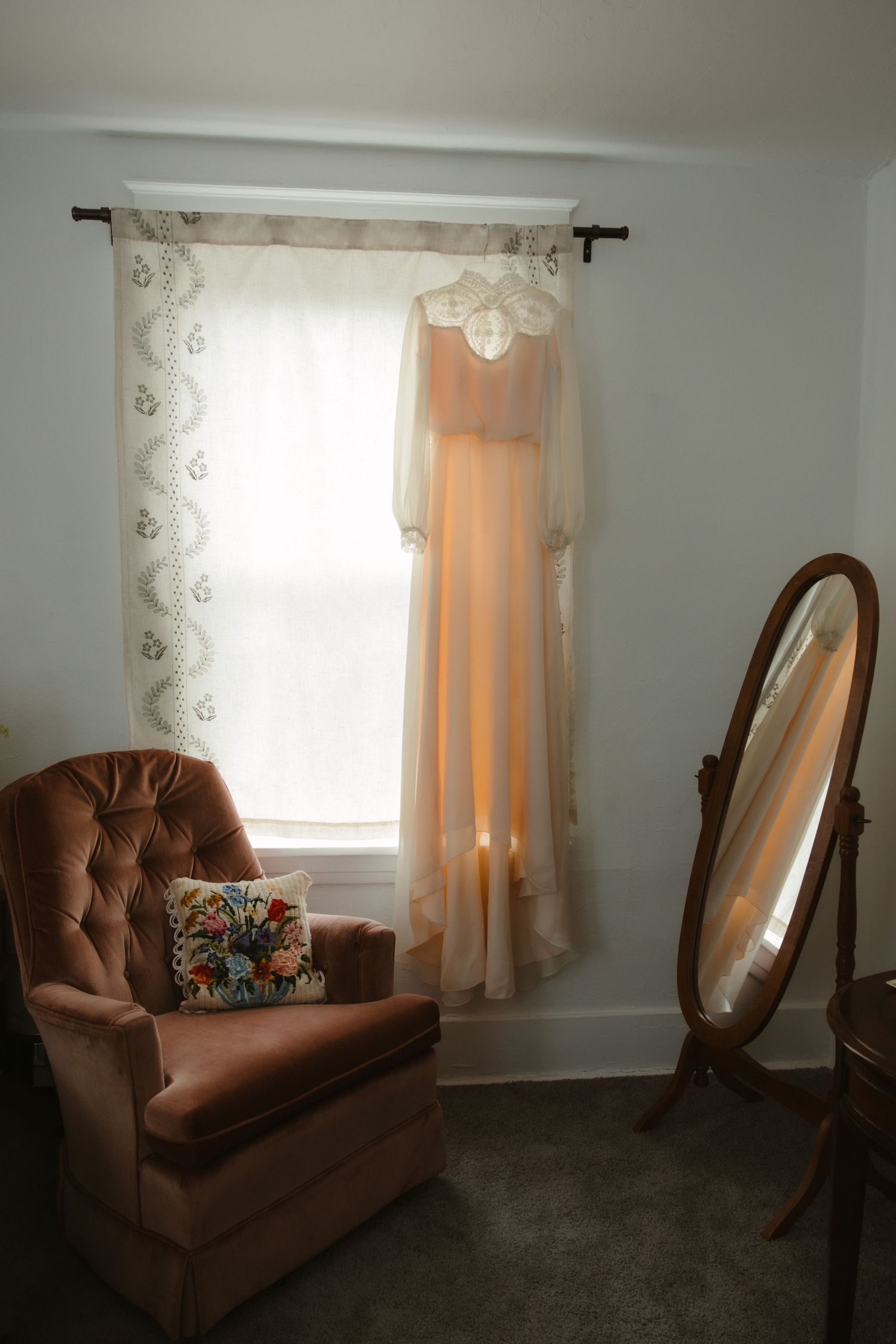 Vintage peach wedding dress hanging in sunlit window beside tufted velvet chair and standing mirror in cozy bedroom.