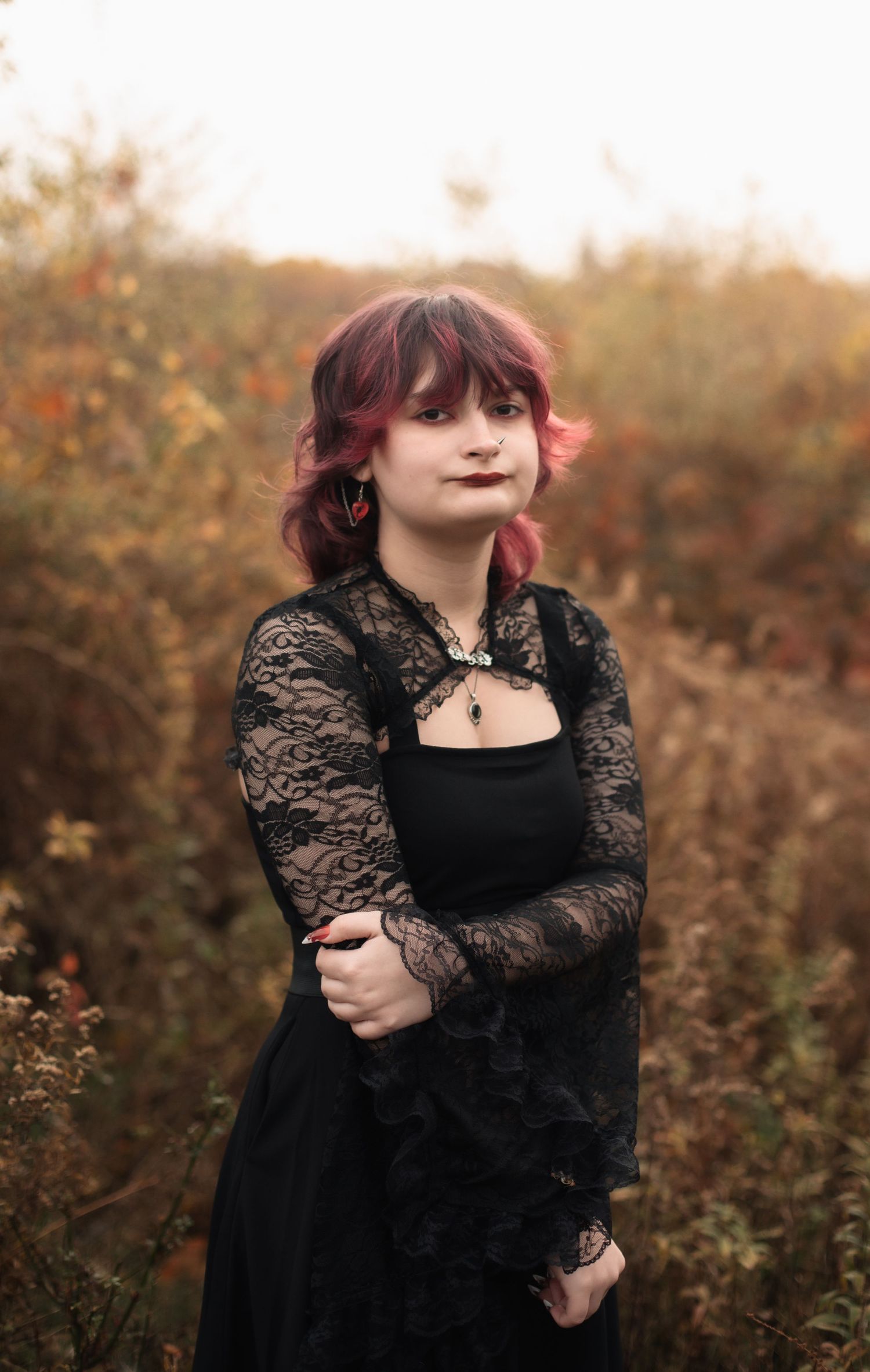 A person in a black lace dress poses thoughtfully in an autumn field at golden hour.