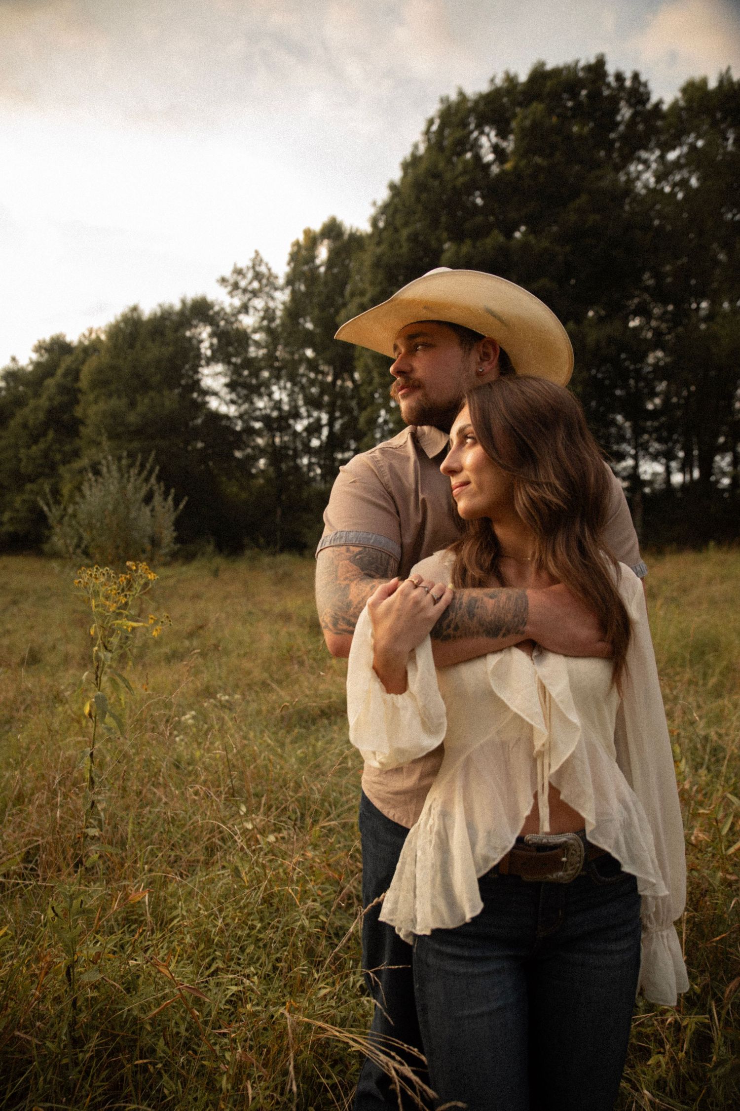 A romantic couple embraces in a golden field at sunset wearing western attire and cowboy hats.