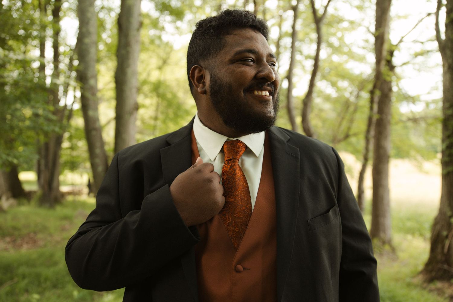Smiling man in dark suit with orange vest and tie standing outdoors among trees.