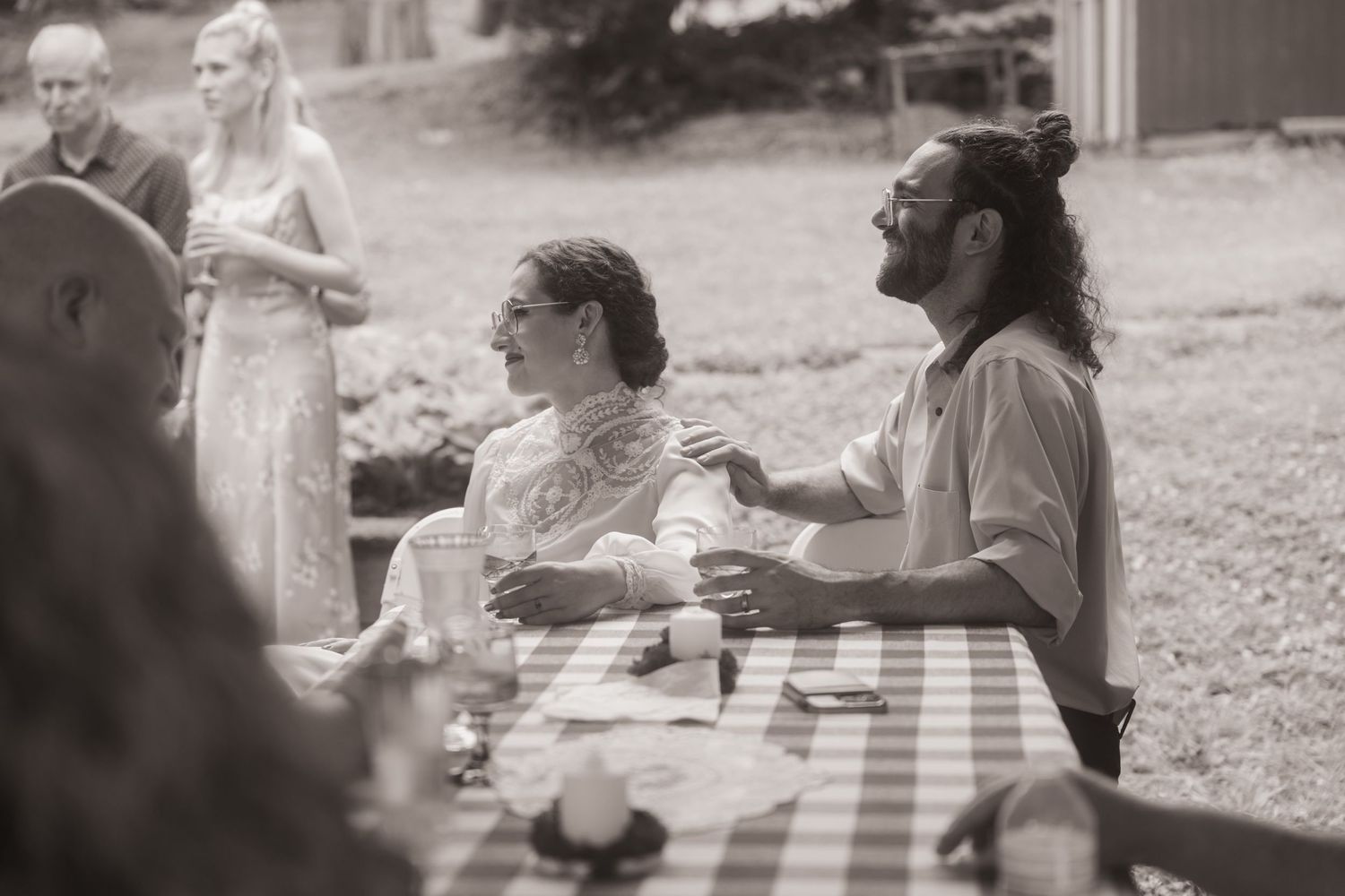 Black and white photo of a couple at a checkered tablecloth outdoor wedding reception, sharing a joyful moment.