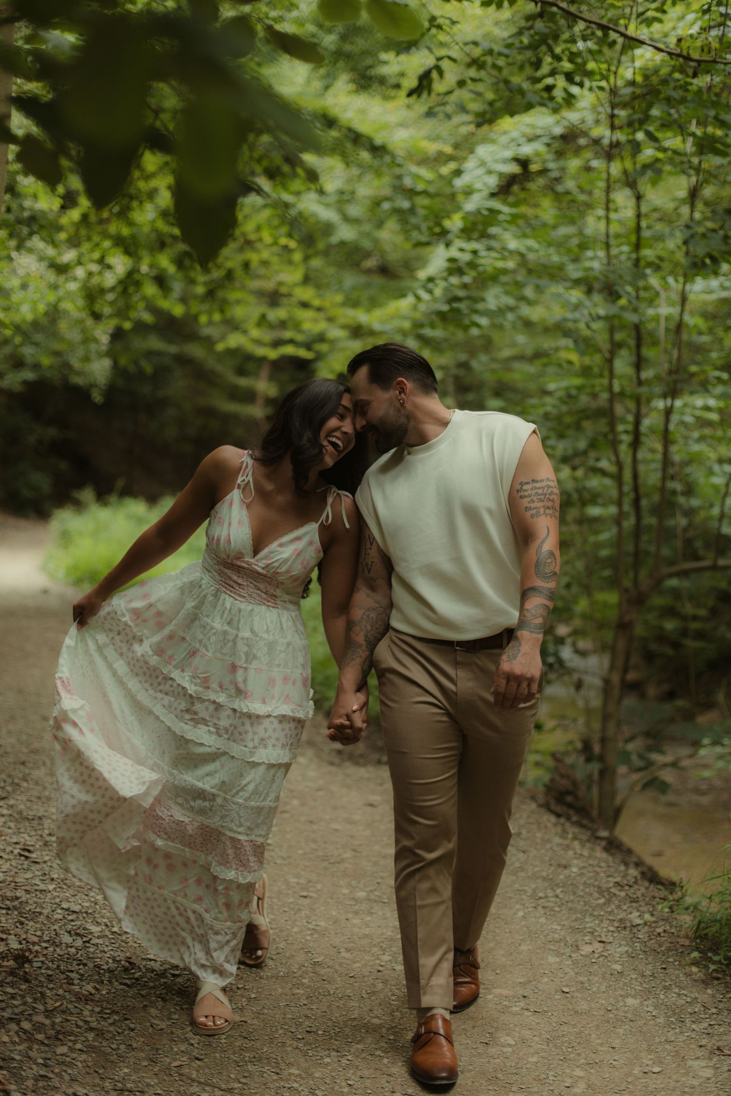 Couple walking hand-in-hand on a forest path, laughing together, woman in white lace dress, man in beige outfit with tattoos.