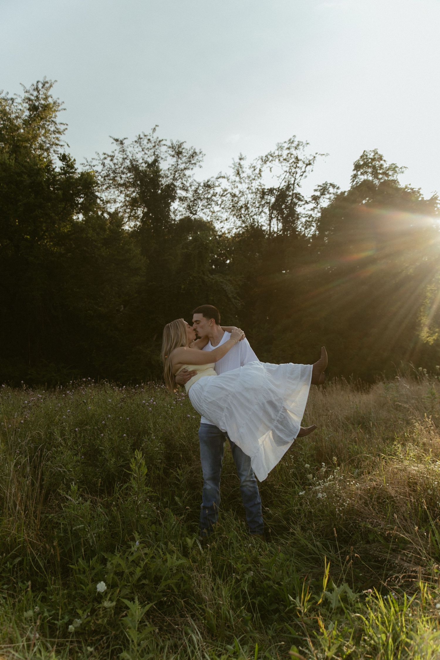 A couple shares a romantic kiss in a sunlit meadow, the man lifting the woman in a white dress at golden hour.
