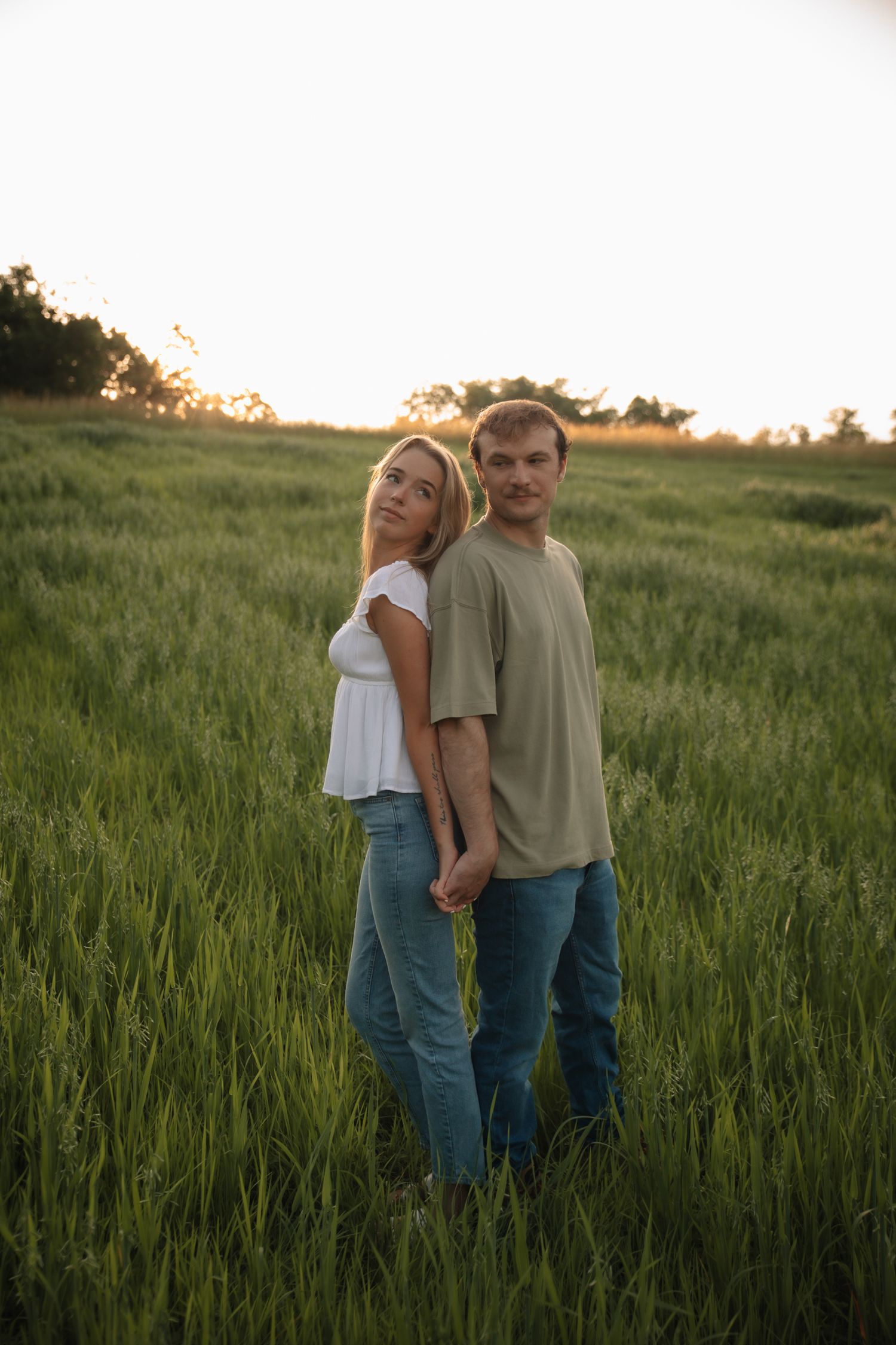 A couple stands together in a lush green field during golden hour as the sun sets in the distance.