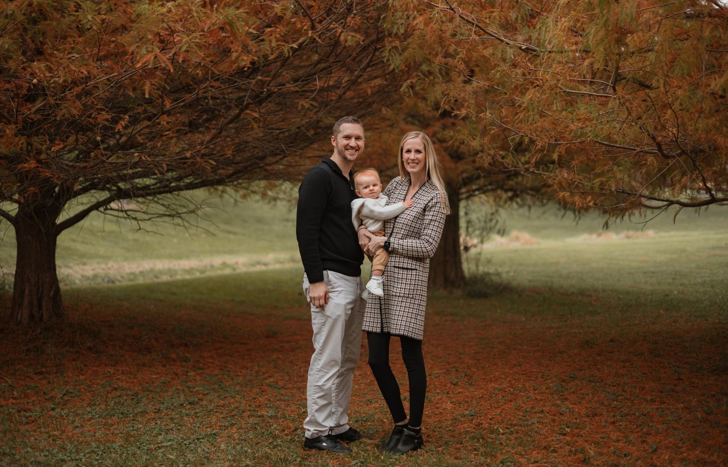 A family poses together under a large autumn tree with orange foliage on a carpet of fallen leaves.
