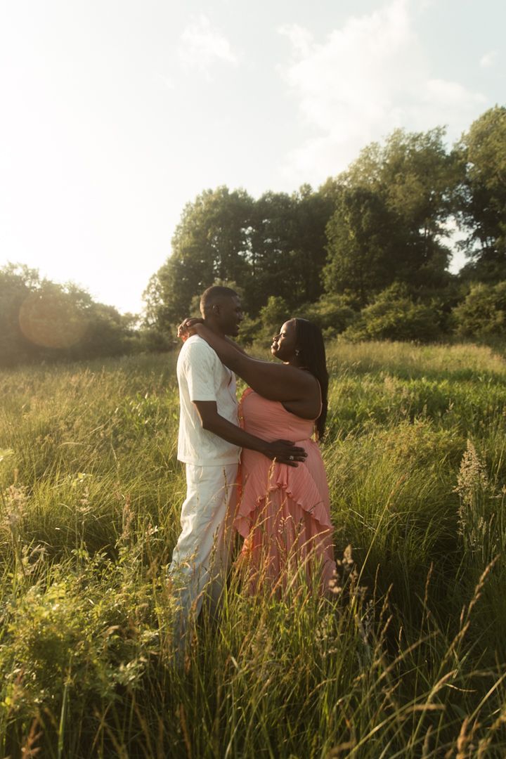 A couple embraces in a sunlit meadow, the woman in a pink dress, surrounded by lush green grass and trees.