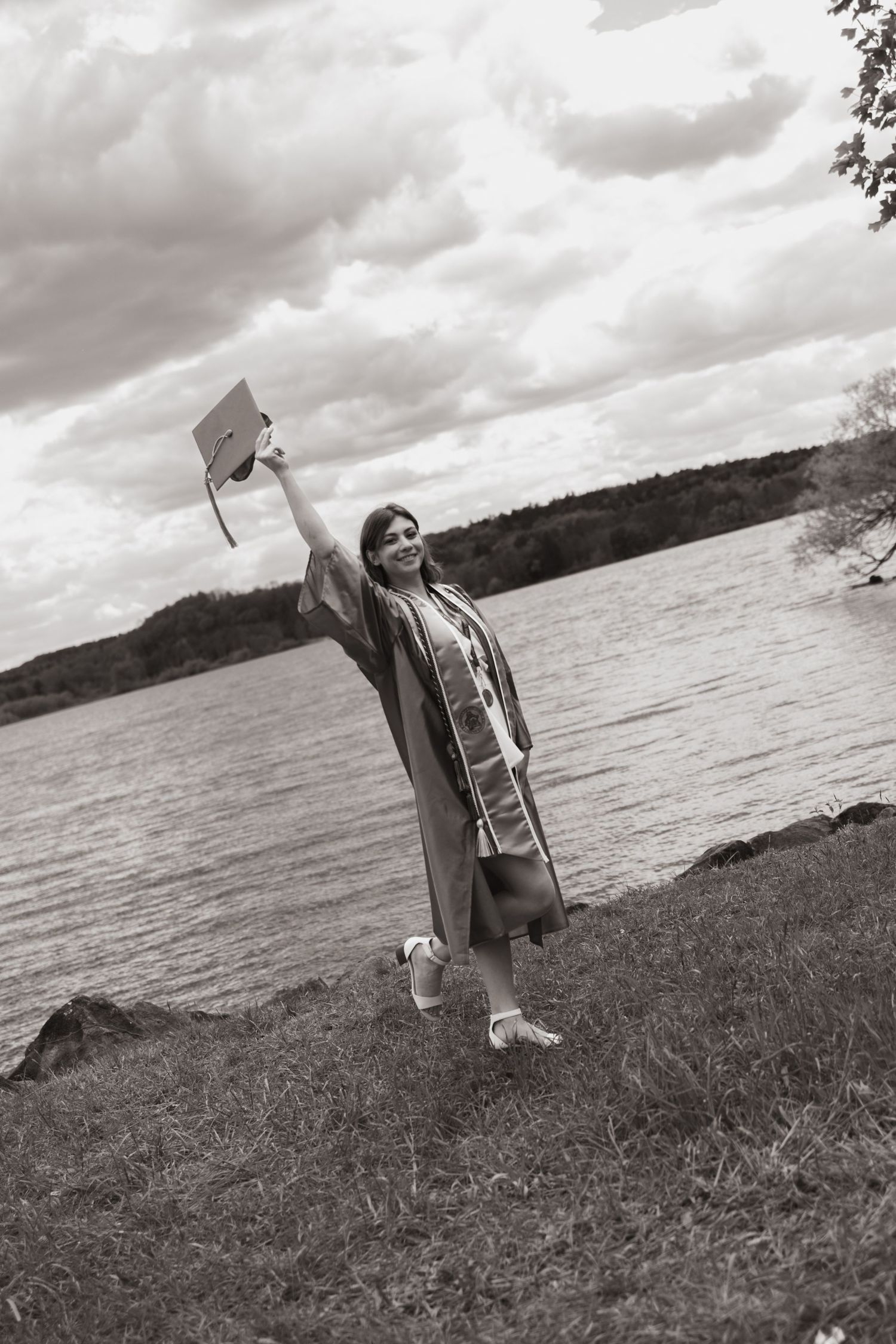 Black and white photo of a smiling graduate in cap and gown raising her mortarboard by a scenic lakeside.