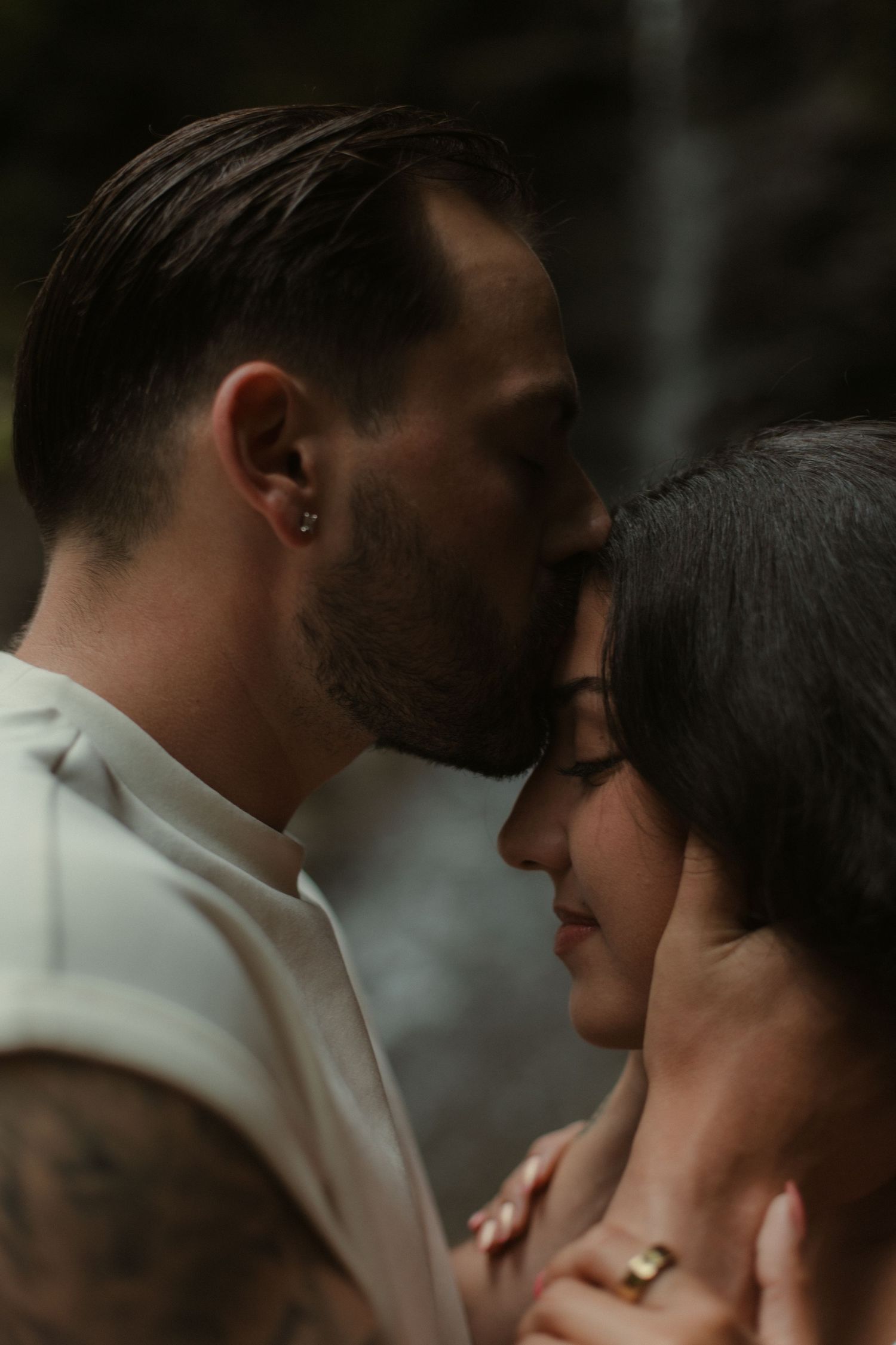 A man with tattoos kisses a woman's forehead tenderly, both wearing rings in a romantic, intimate moment.