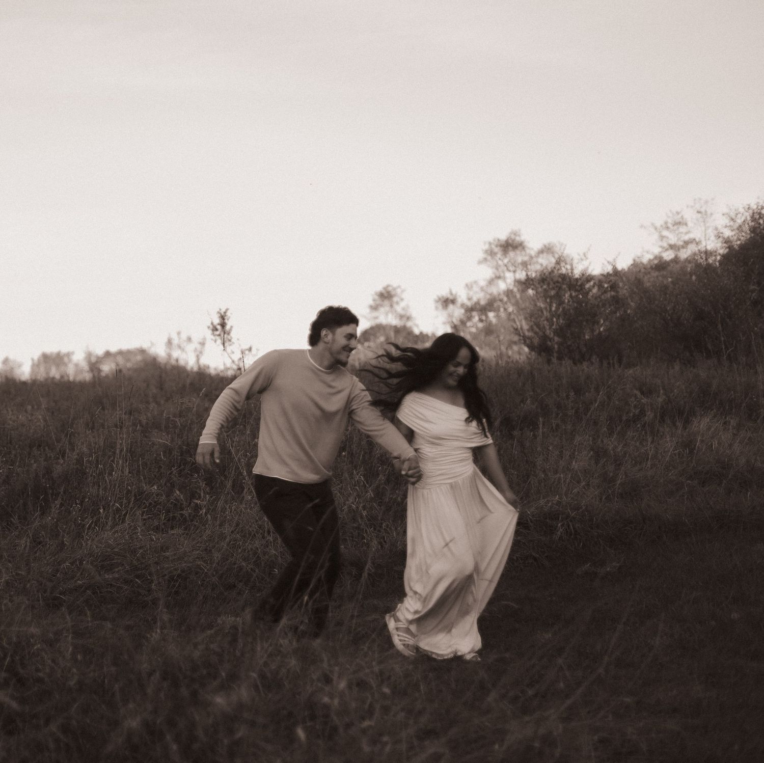 A couple joyfully runs through a grassy field in a sepia-toned romantic outdoor photo session.