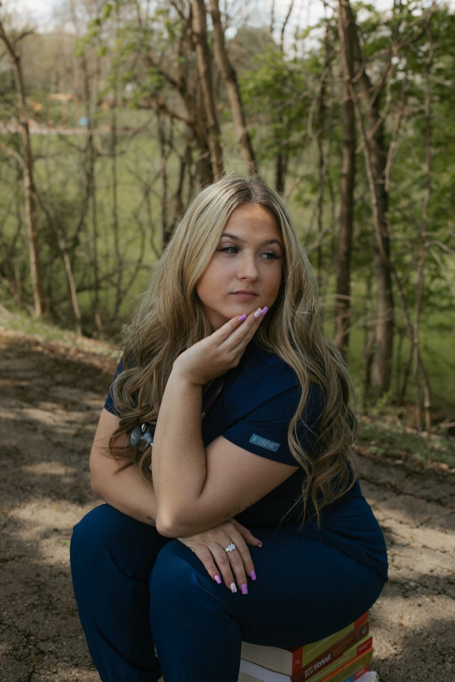 Blonde woman in navy blue scrubs sitting outdoors on a wooded trail, posing thoughtfully with hand on cheek.