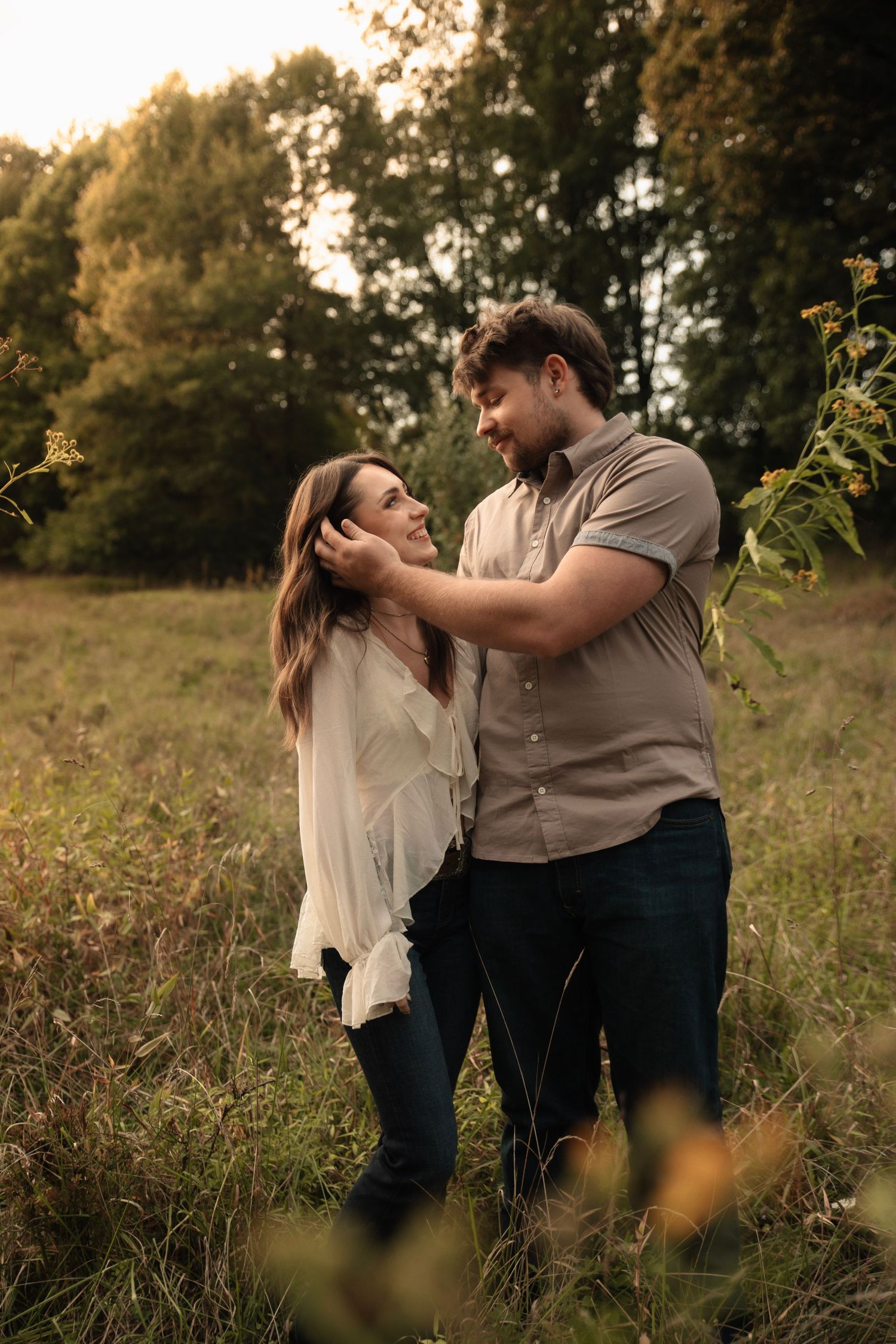 A couple shares a tender moment in a sunlit meadow, the man gently touching the woman's hair as they gaze at each other.