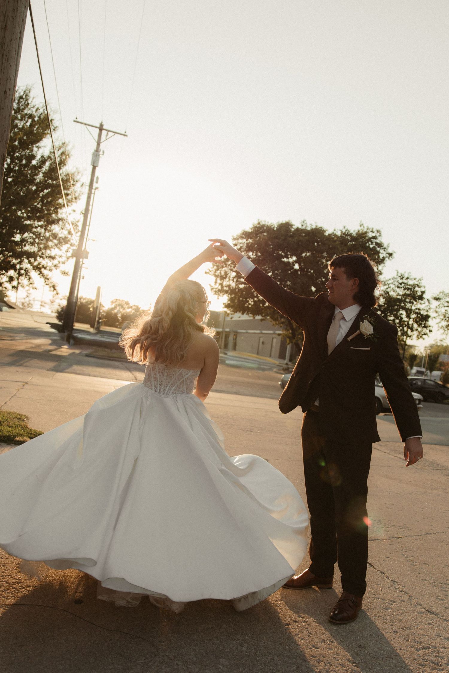 Groom twirling bride in white ball gown on sunlit street during golden hour wedding portrait.