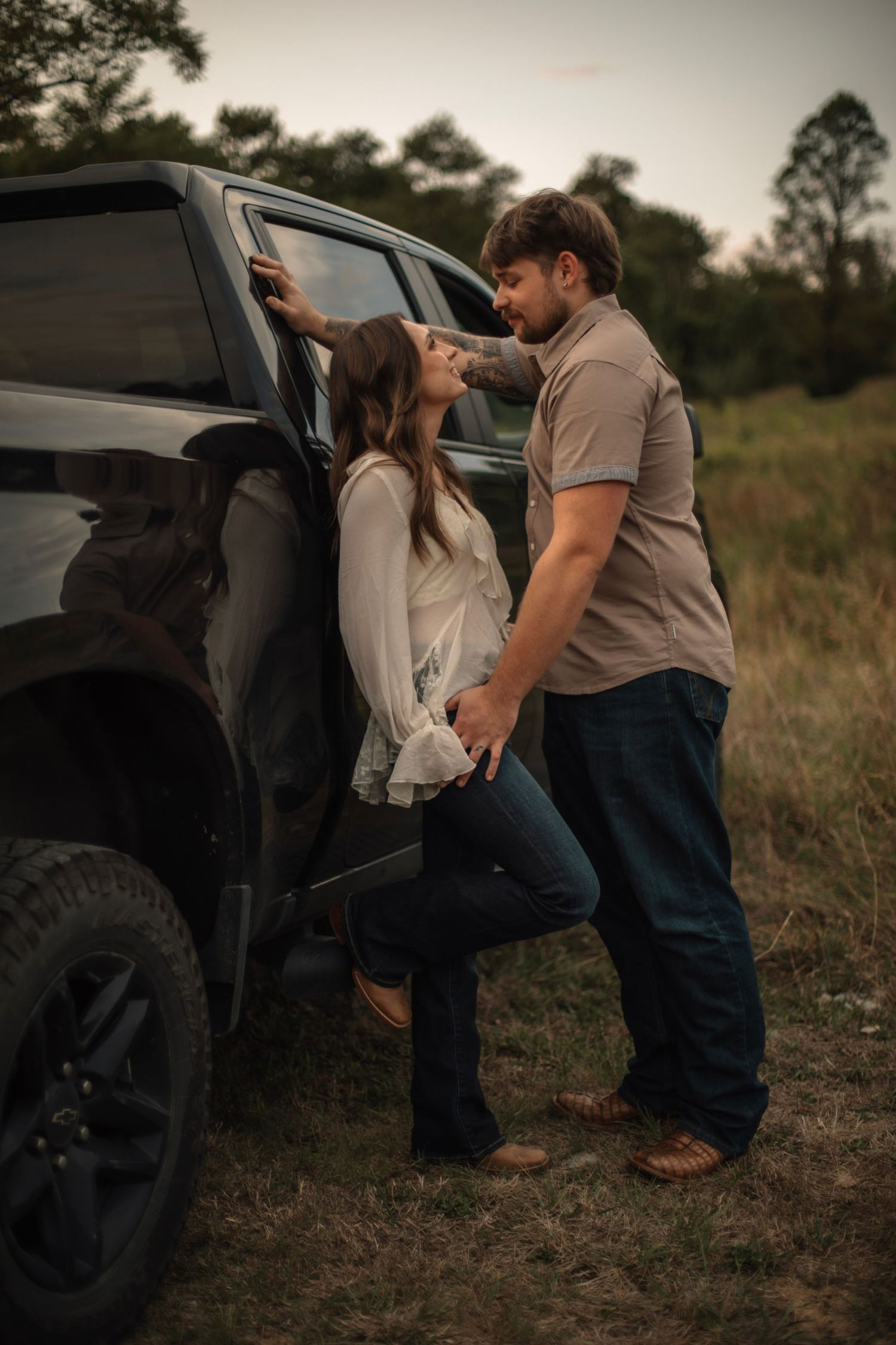 Romantic couple sharing a tender moment leaning against a black SUV in a rural field at dusk.