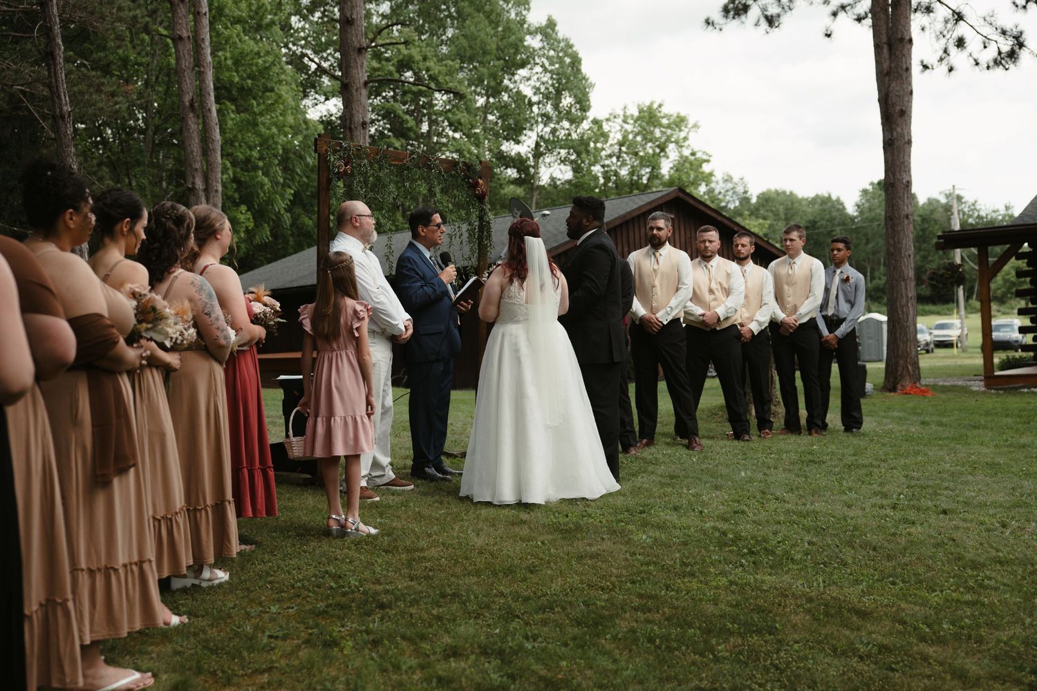 Outdoor wedding ceremony on green lawn with bridesmaids in dusty rose dresses and groomsmen in black suits under rustic wooden arch.