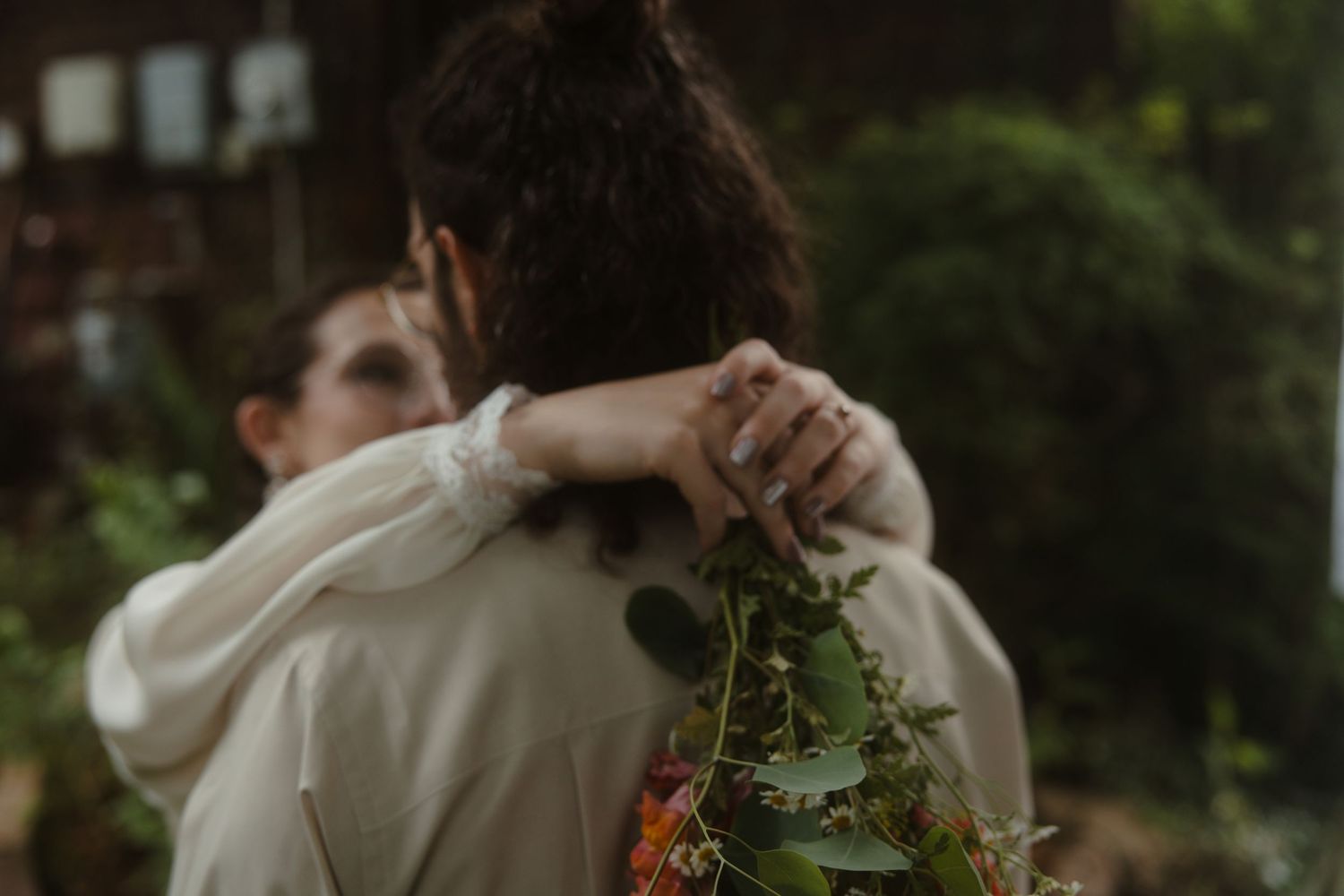 Two people embracing outdoors, one holding a bouquet of greenery, wearing a ring, in a lush garden setting.