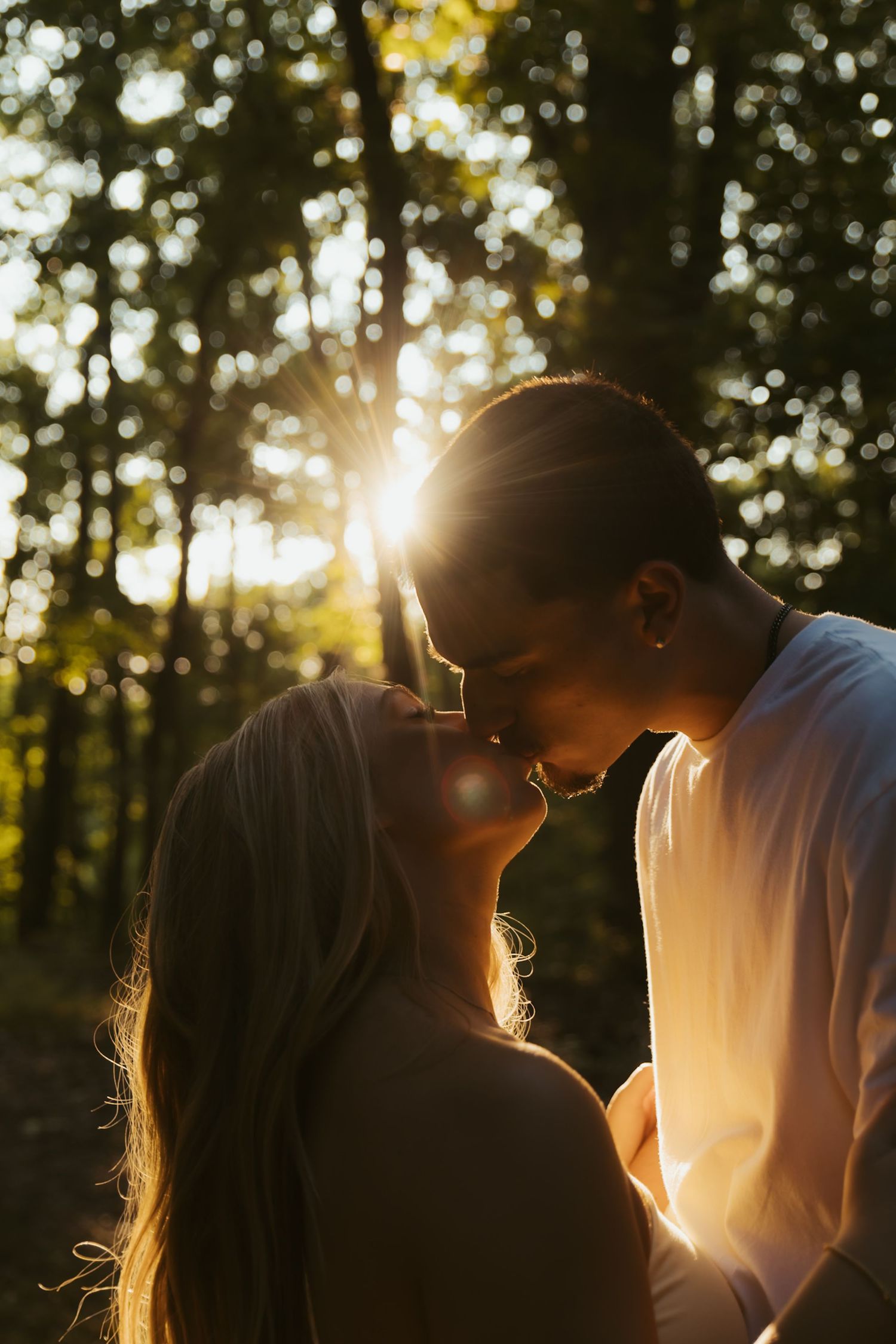 Romantic couple sharing a kiss in a sunlit forest with golden backlight shining through the trees.
