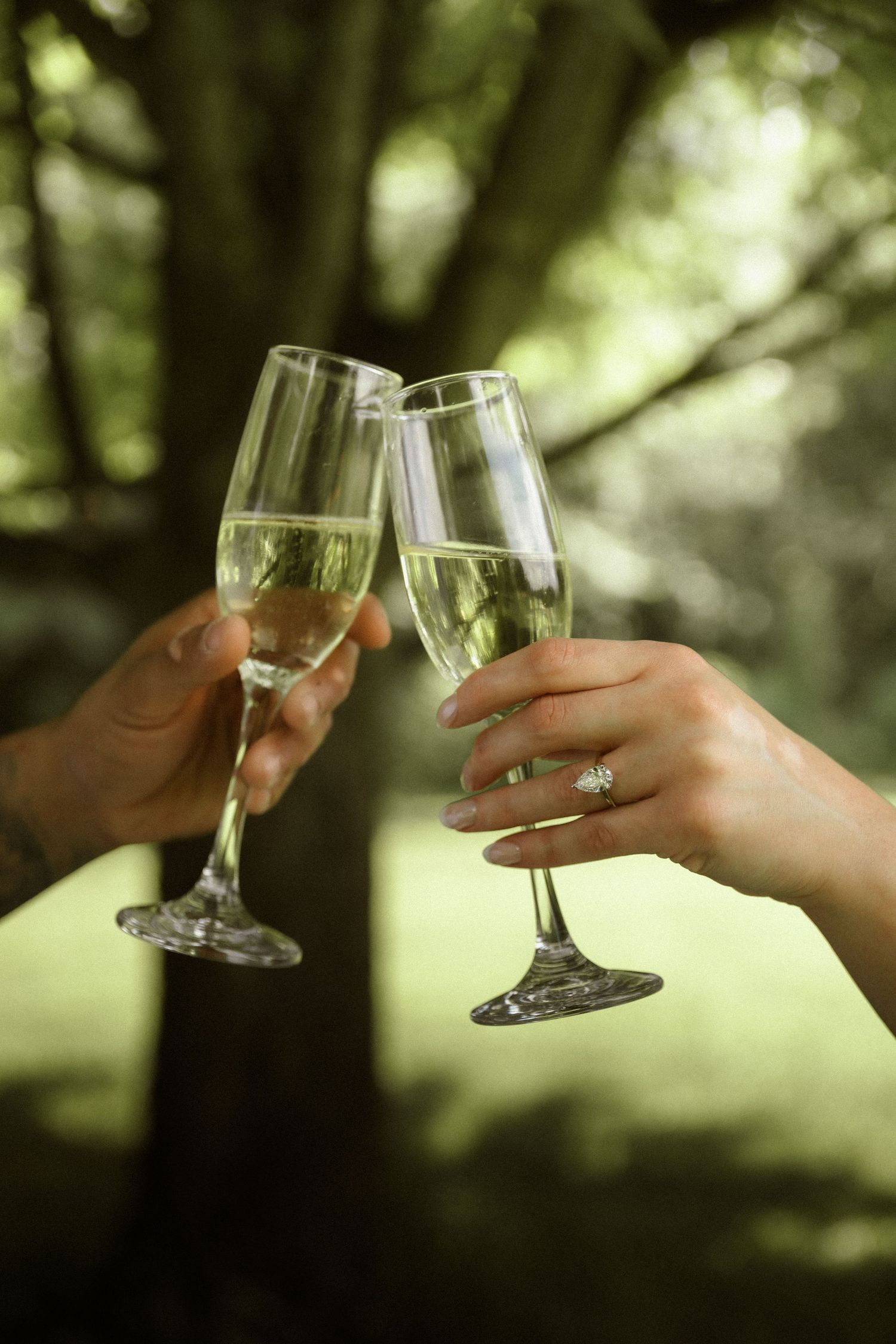 Two hands clinking champagne flutes outdoors, one adorned with an engagement ring, celebrating a special occasion.