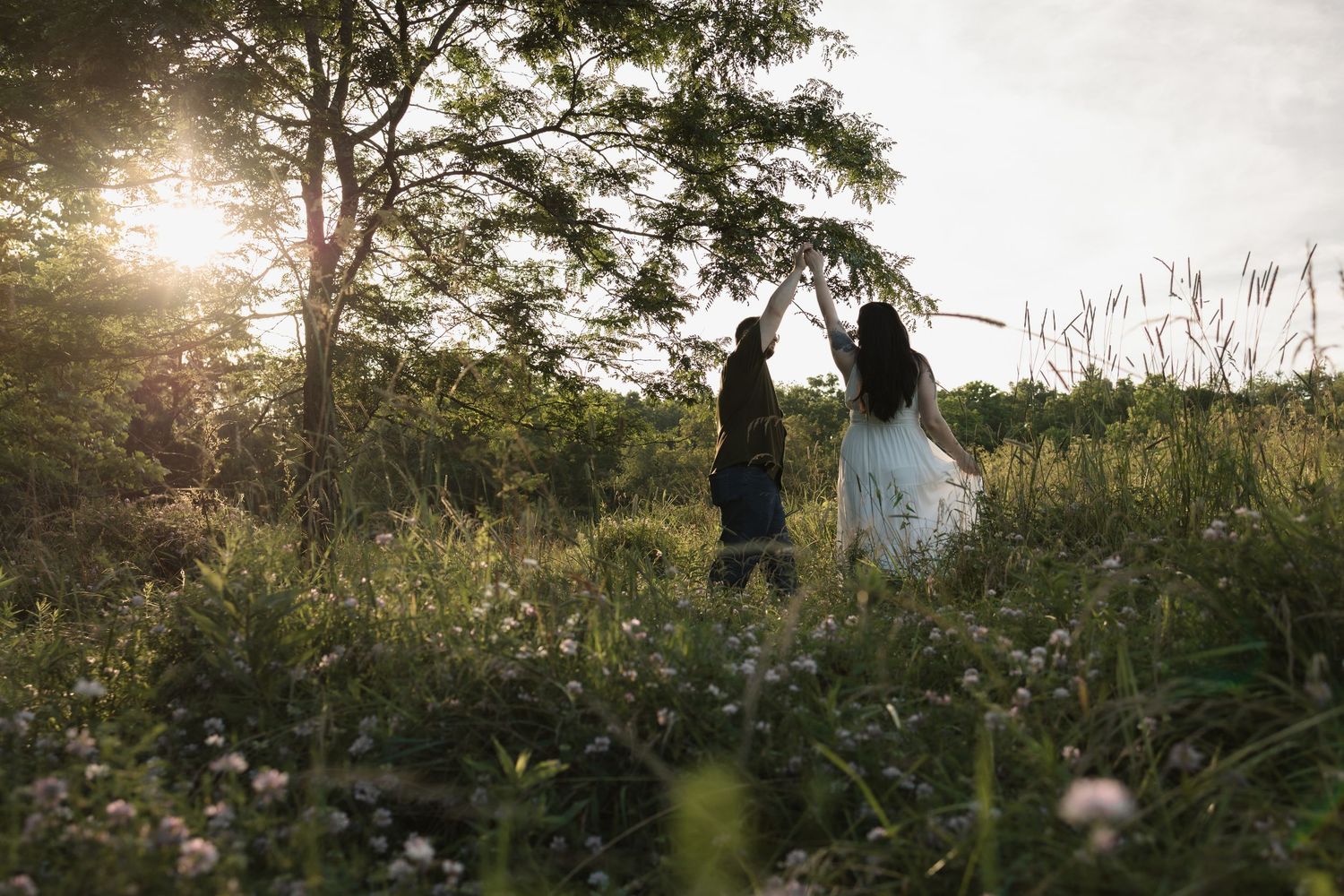 Couple dancing in a sunlit meadow surrounded by wildflowers and trees at golden hour.