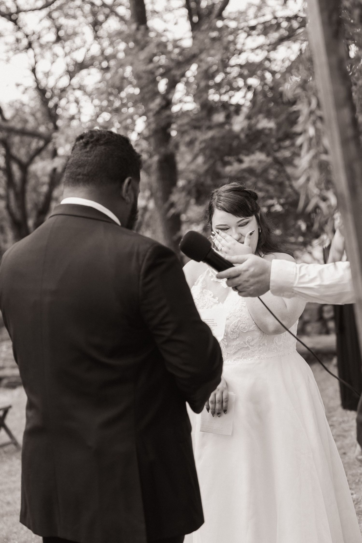 Black and white photo of emotional bride wiping tears during outdoor wedding ceremony while groom reads vows.