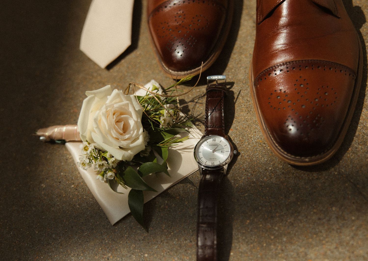 Brown leather dress shoes, a boutonniere with white roses, and a brown leather watch arranged on a surface.
