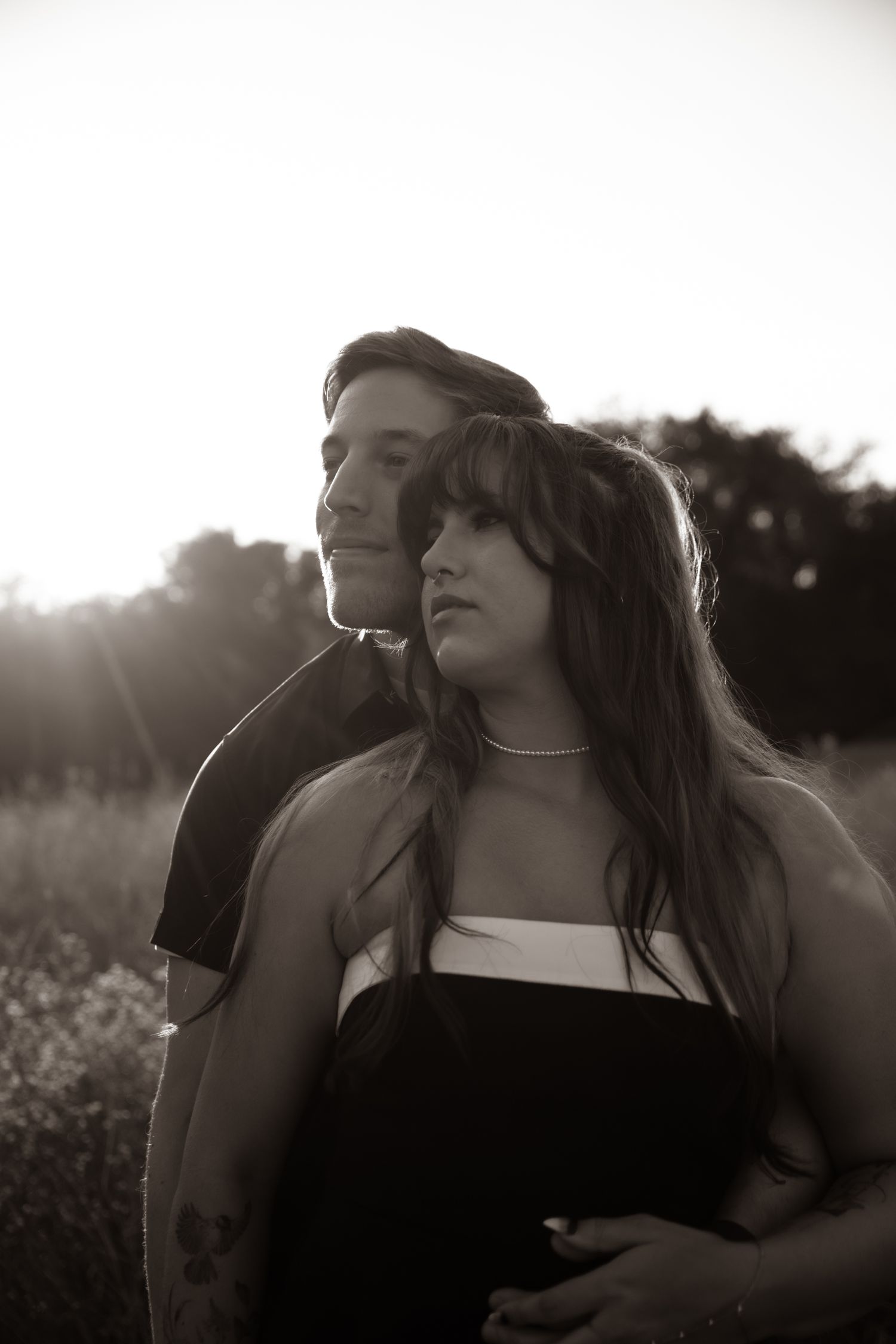 Black and white photo of a young couple embracing outdoors at sunset, woman in strapless dress with long hair.