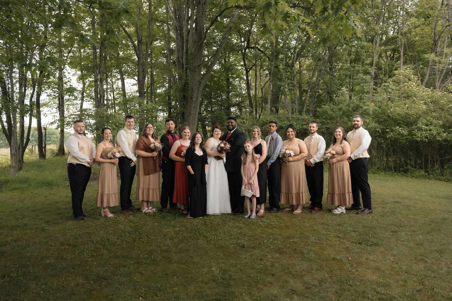 Wedding party posing outdoors on green lawn surrounded by tall trees, bridesmaids in earth tones flanking bride and groom.