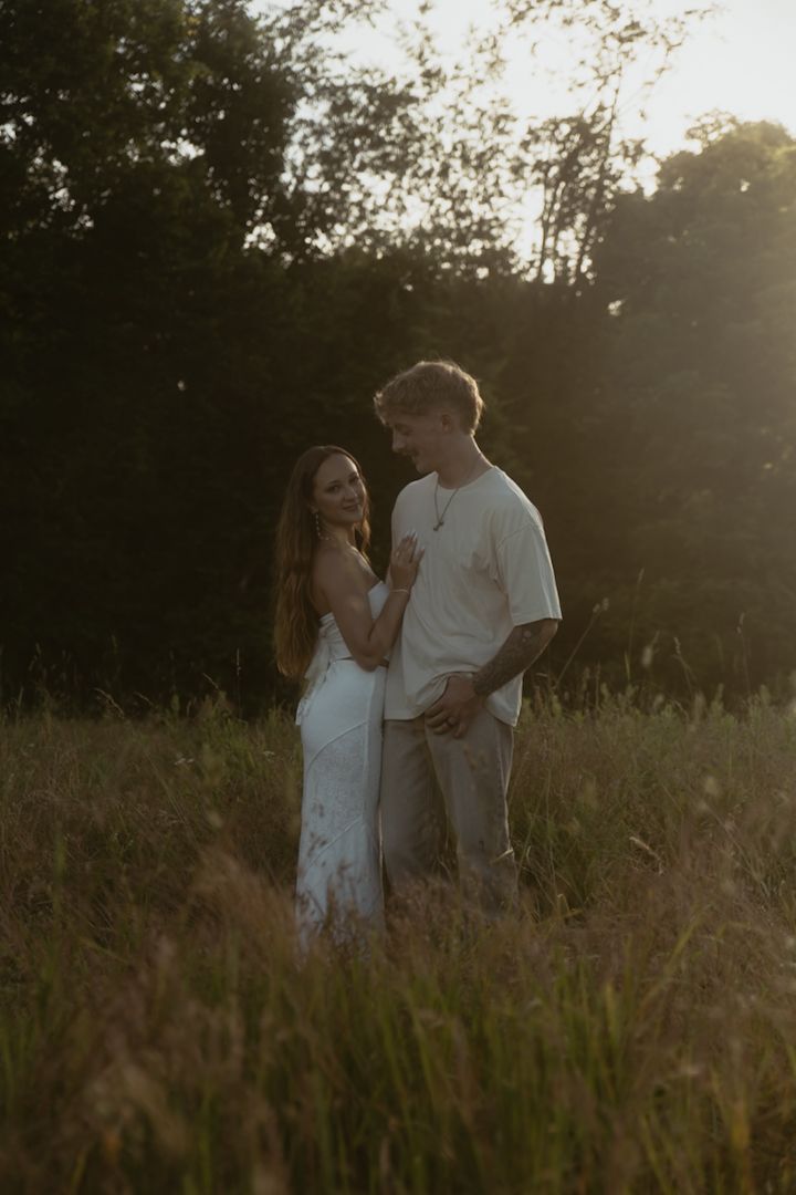 Couple embracing in golden hour sunlight in a meadow, woman in white dress and man in white shirt with tattoos.