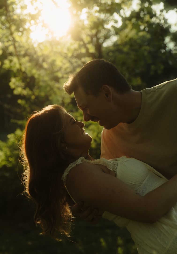 A couple shares a romantic almost-kiss moment outdoors, bathed in warm golden sunlight filtering through lush green trees.